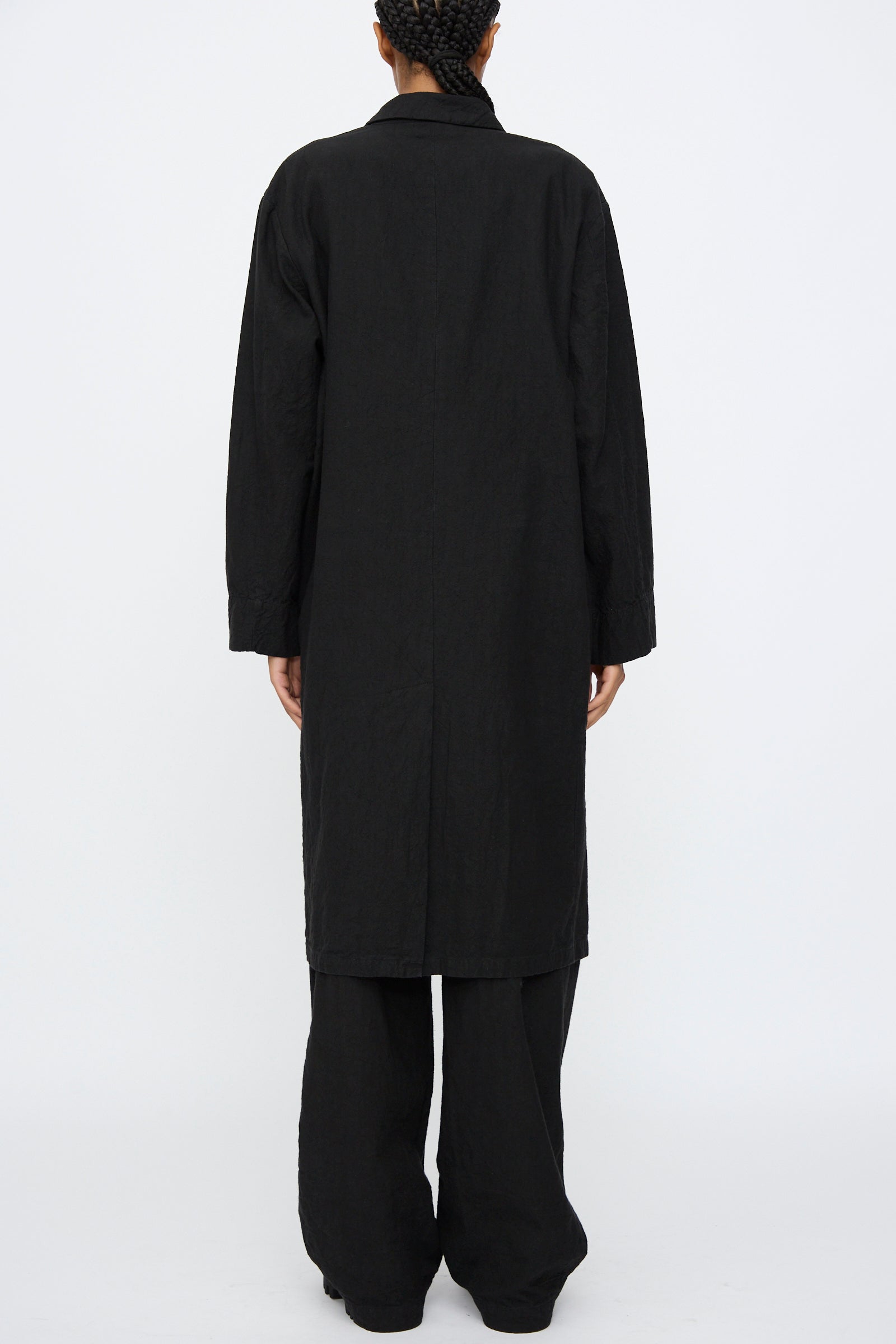 A person stands facing away from the camera in Black Crane's Crinkled Coat in Black, featuring a subtle crinkled texture, paired with black pants against a plain white background.