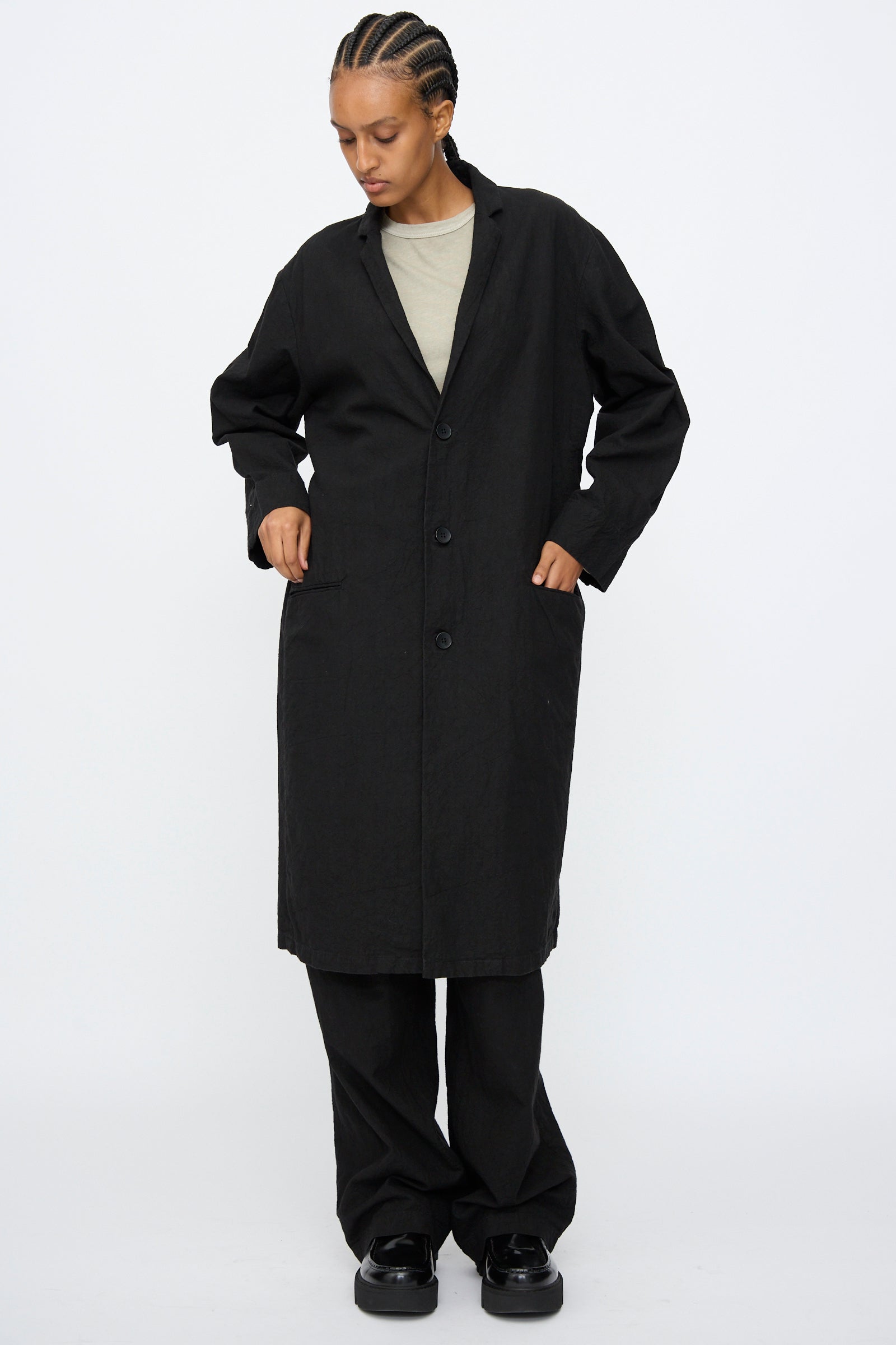 A person wears the Black Crane Crinkled Coat in Black over a light beige top, black pants, and black shoes, standing against a plain white background.