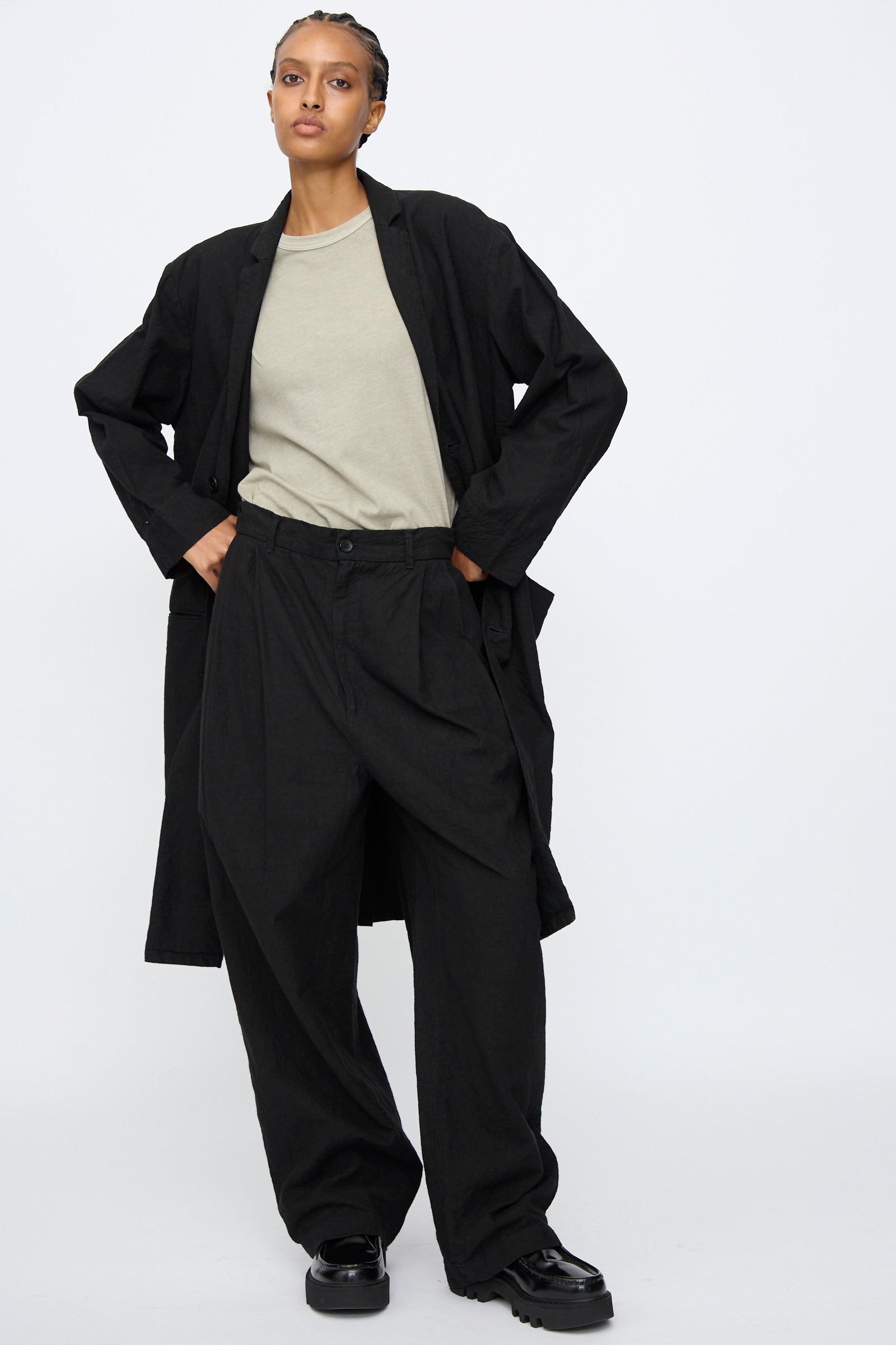 A person wearing a light gray t-shirt, Black Crane Crinkled Coat in Black, black wide-leg pants, and black shoes stands against a plain white background with their hands in their pockets.