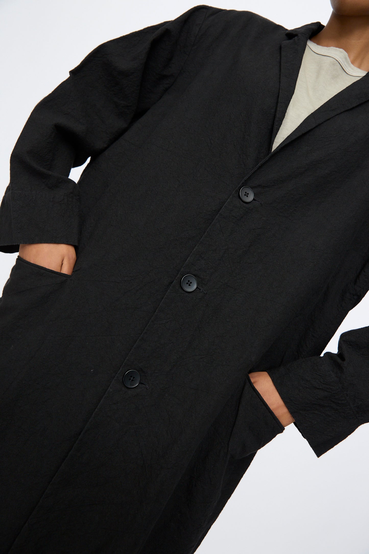 A person wears Black Crane’s Crinkled Coat in Black— a long Japanese linen coat with buttons, side pockets, and a textured finish—layered over a light top against a plain background.