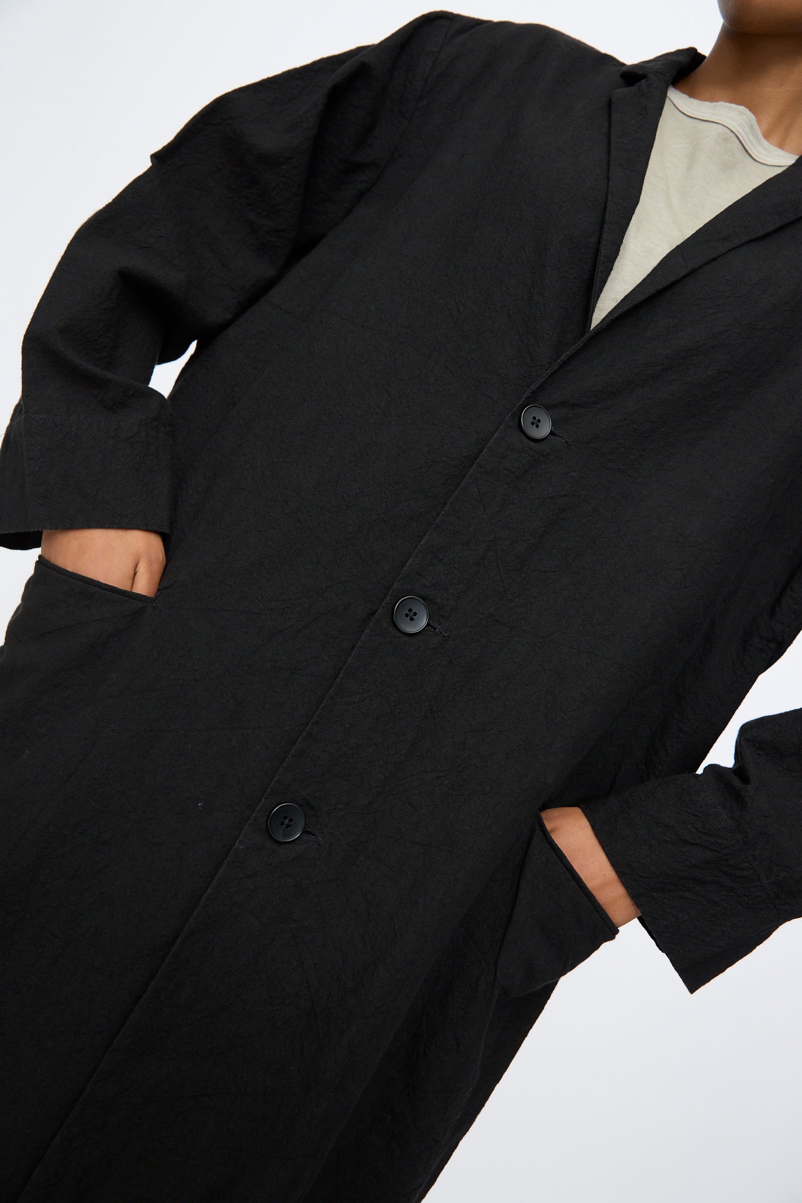 A person wears Black Crane’s Crinkled Coat in Black— a long Japanese linen coat with buttons, side pockets, and a textured finish—layered over a light top against a plain background.