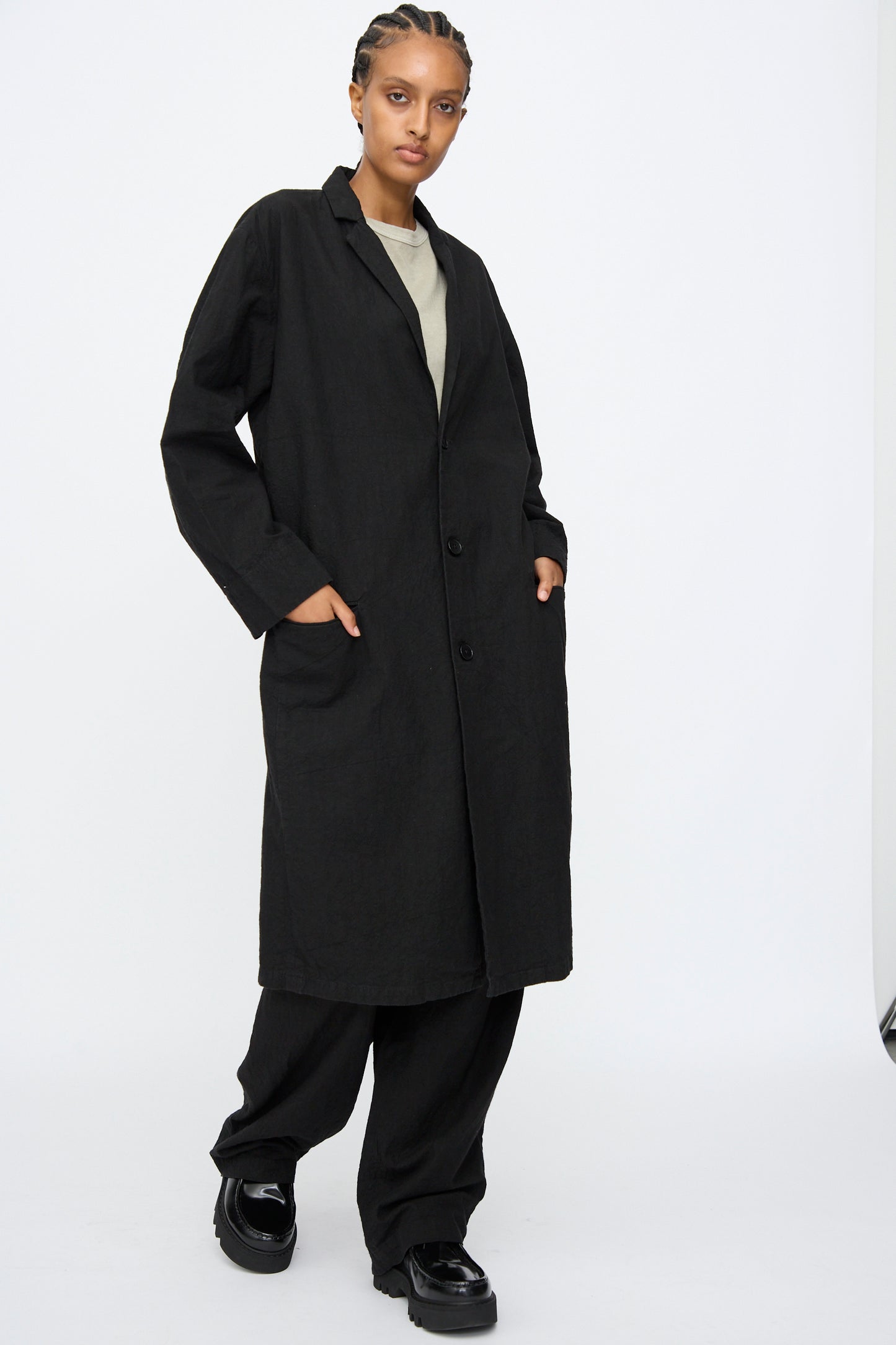 A person stands against a white background wearing the Black Crane Crinkled Coat in Black, paired with loose black pants, black shoes, and a light-colored top.