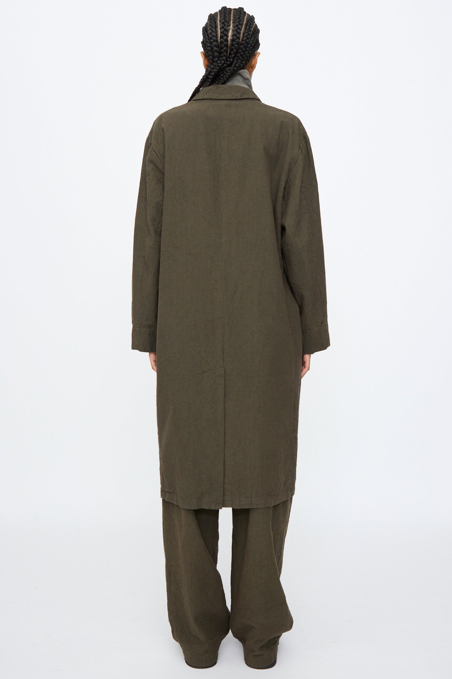 Person standing with back to camera, wearing the Black Crane Crinkled Coat in Charcoal with matching wide-leg pants, set against a plain white background.