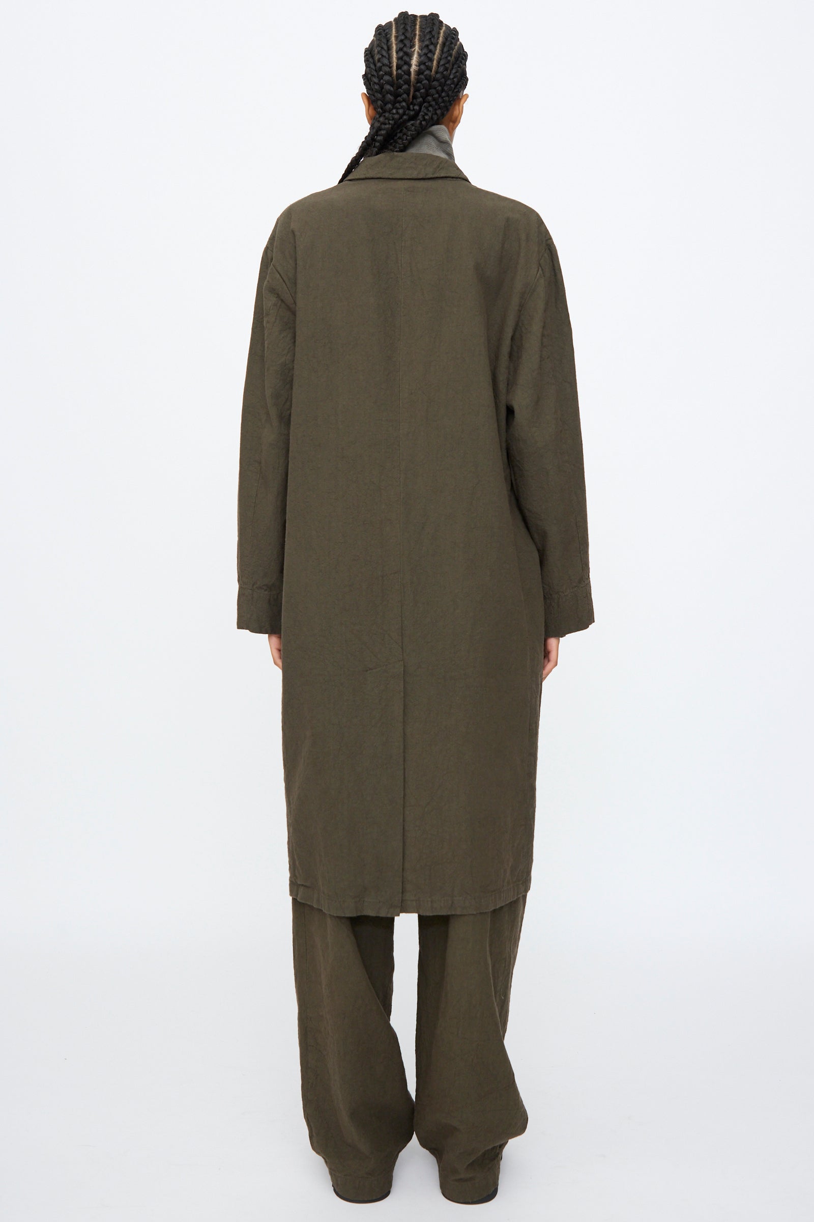 Person standing with back to camera, wearing the Black Crane Crinkled Coat in Charcoal with matching wide-leg pants, set against a plain white background.