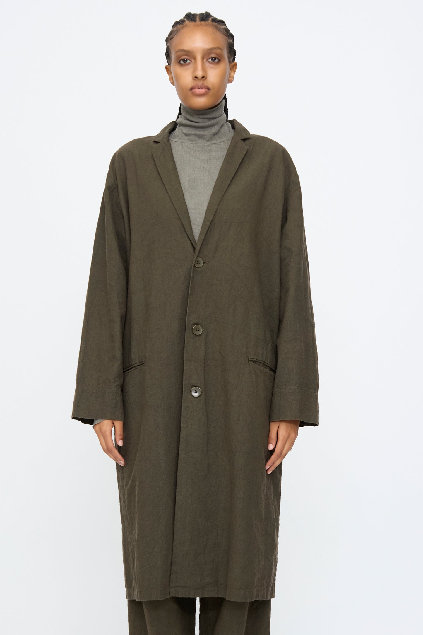 A person wears the Black Crane Crinkled Coat in Charcoal over a gray turtleneck, standing facing the camera against a plain white background.