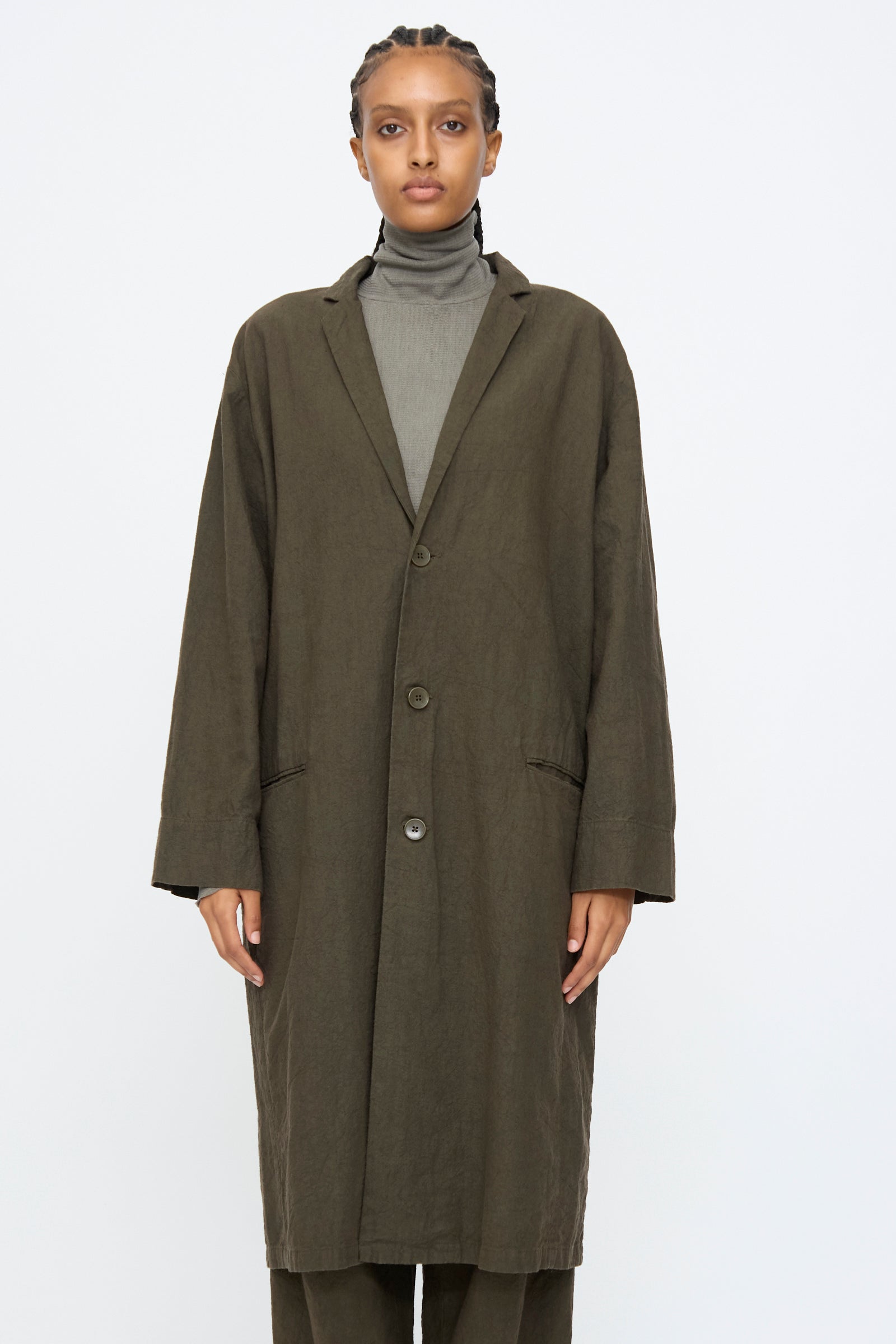 A person wears the Black Crane Crinkled Coat in Charcoal over a gray turtleneck, standing facing the camera against a plain white background.