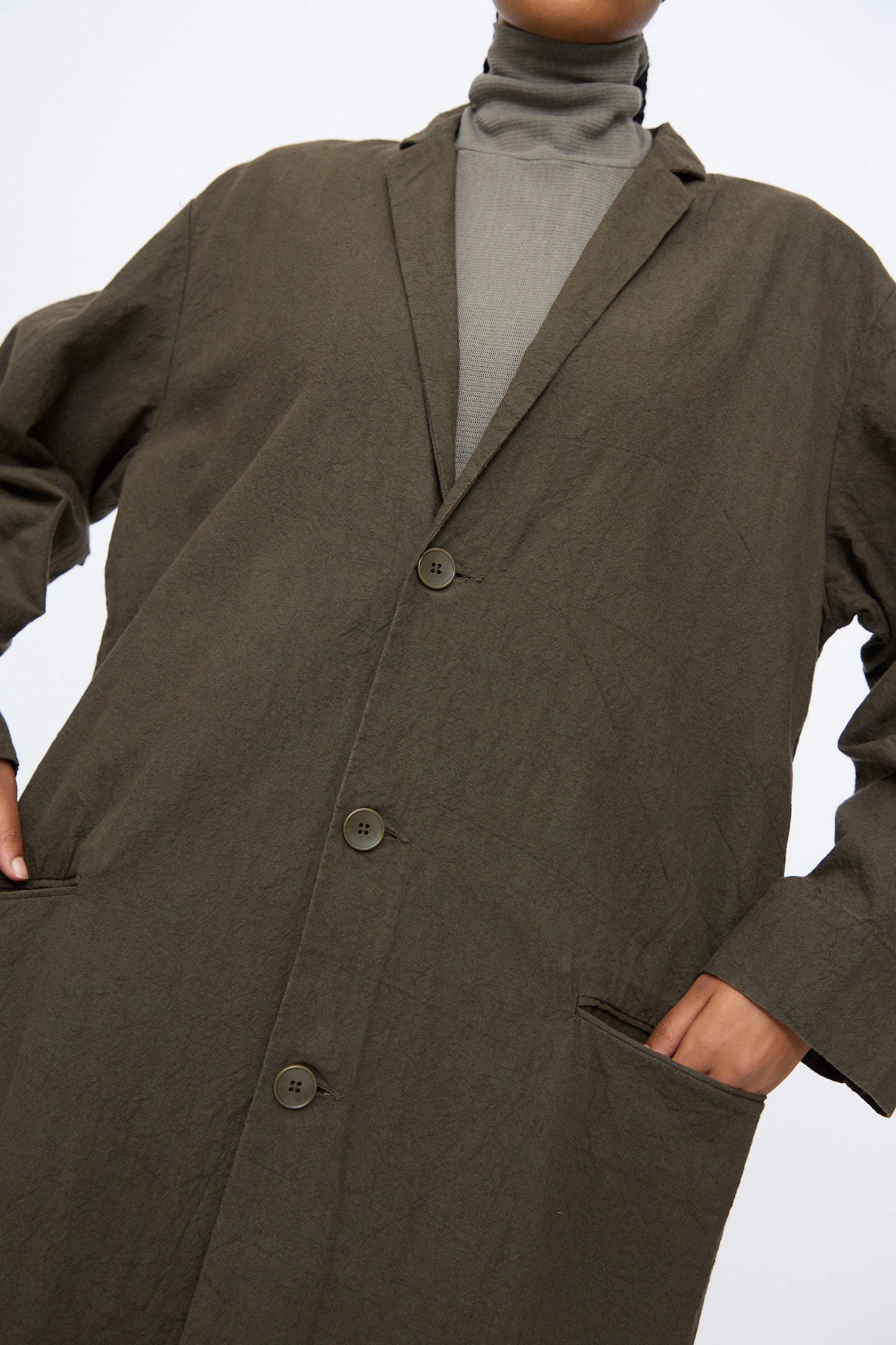 Model wearing the Black Crane Crinkled Coat in Charcoal over a gray turtleneck, hands in pockets, shown from shoulders to mid-thigh.