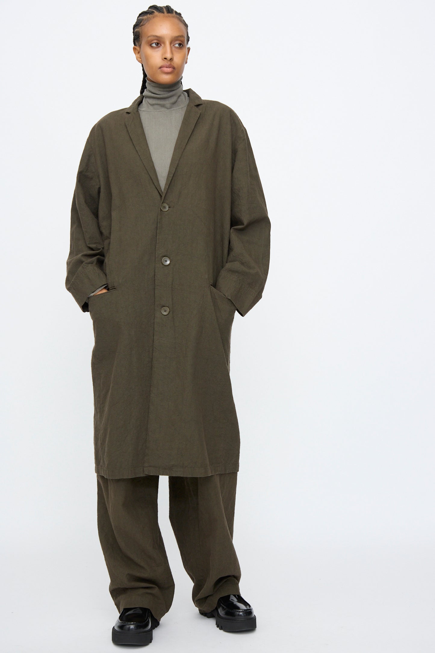 Person stands against a plain background wearing Black Crane’s Crinkled Coat in Charcoal, matching wide-leg pants, a grey turtleneck, and black shoes. Hands are in the coat pockets.