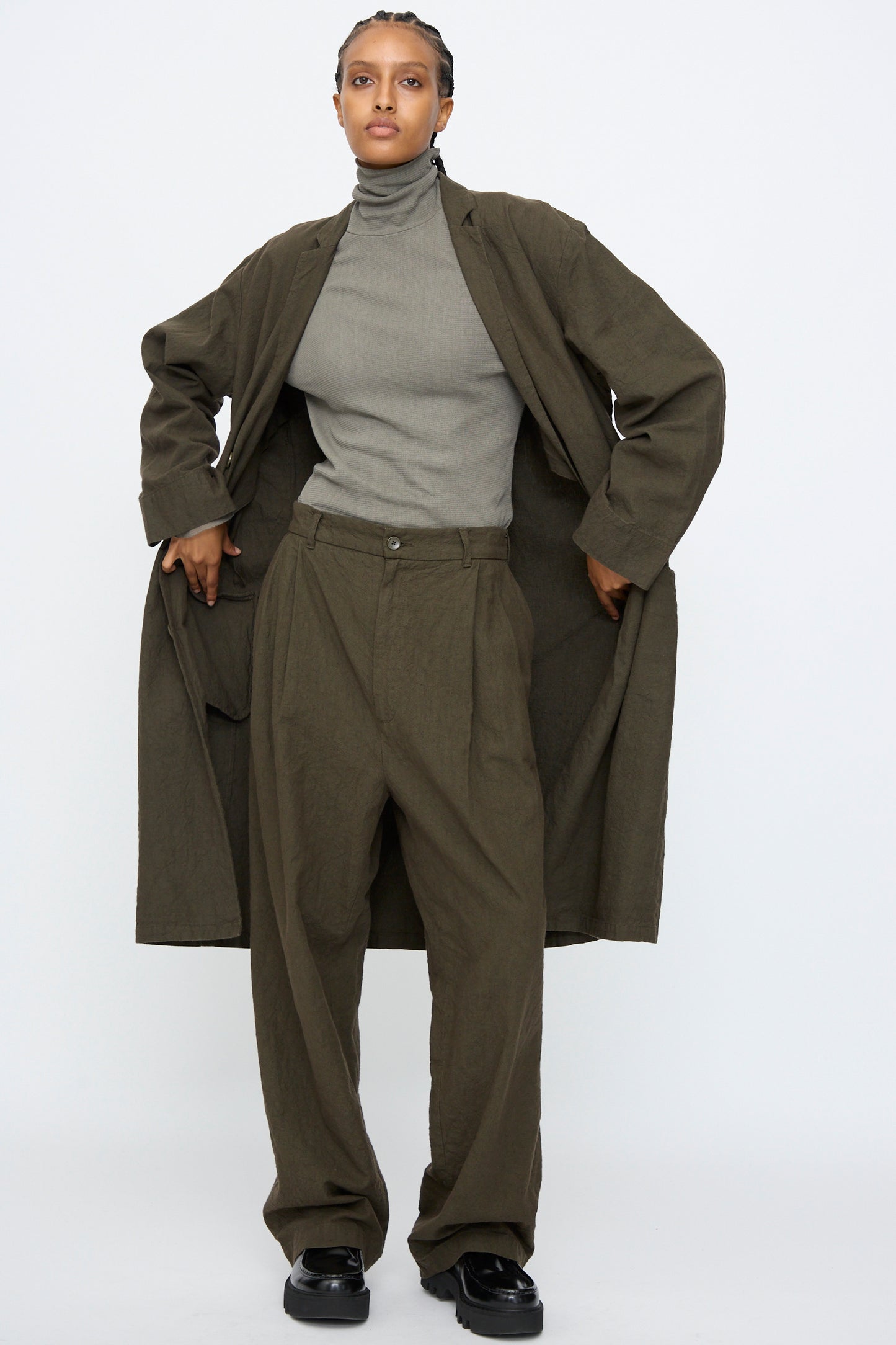 A person wearing Black Crane's Crinkled Coat in Charcoal, a gray turtleneck, loose olive pants, and black shoes stands facing forward against a plain white background.