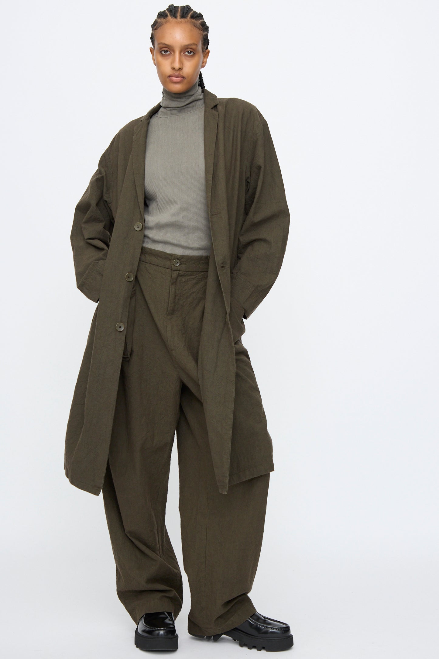 A person wears Black Crane’s Crinkled Coat in Charcoal over a gray turtleneck and an olive green oversized suit with wide-leg pants, standing against a plain white background.