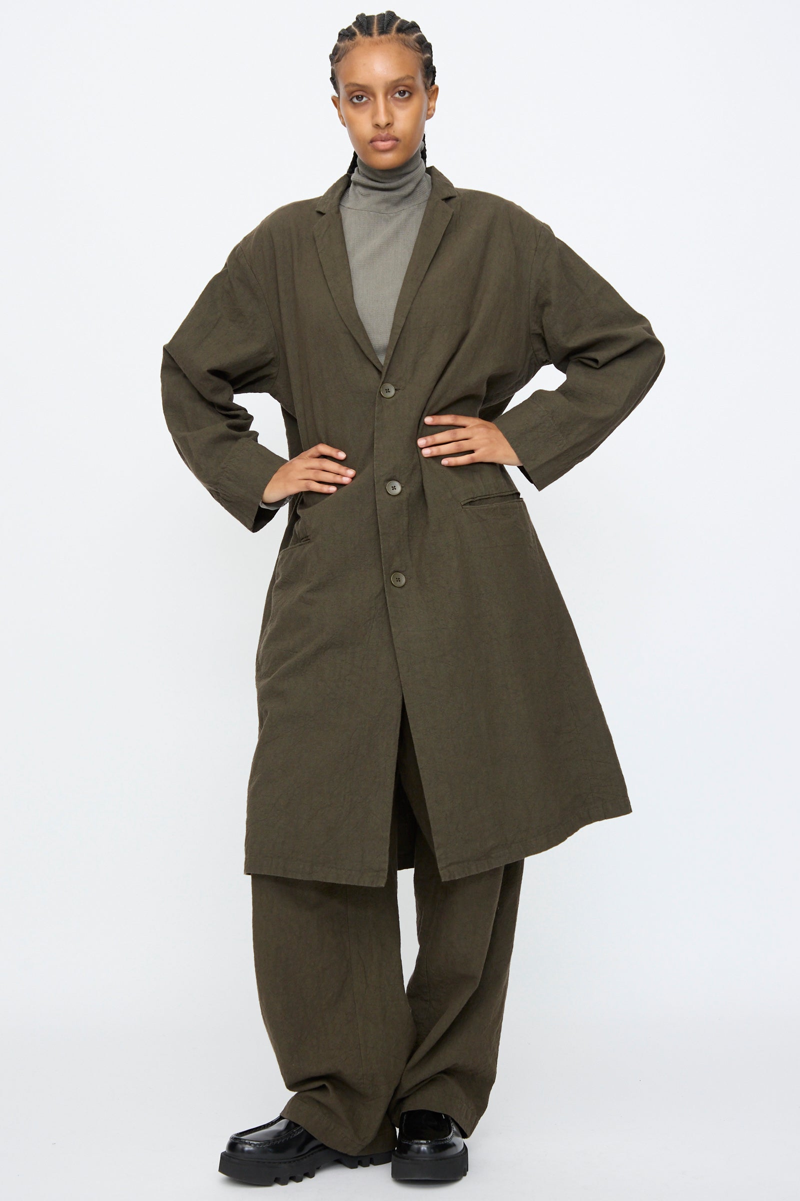 Person wearing the Black Crane Crinkled Coat in Charcoal with matching wide-leg pants, a gray turtleneck, and black shoes, standing against a plain white background.