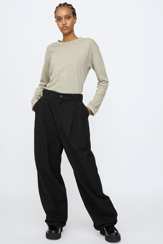 Person with hands in pockets wears a light beige long-sleeve shirt, Black Crane Crinkled Trousers in Black (relaxed fit), and black shoes, standing against a plain white background.