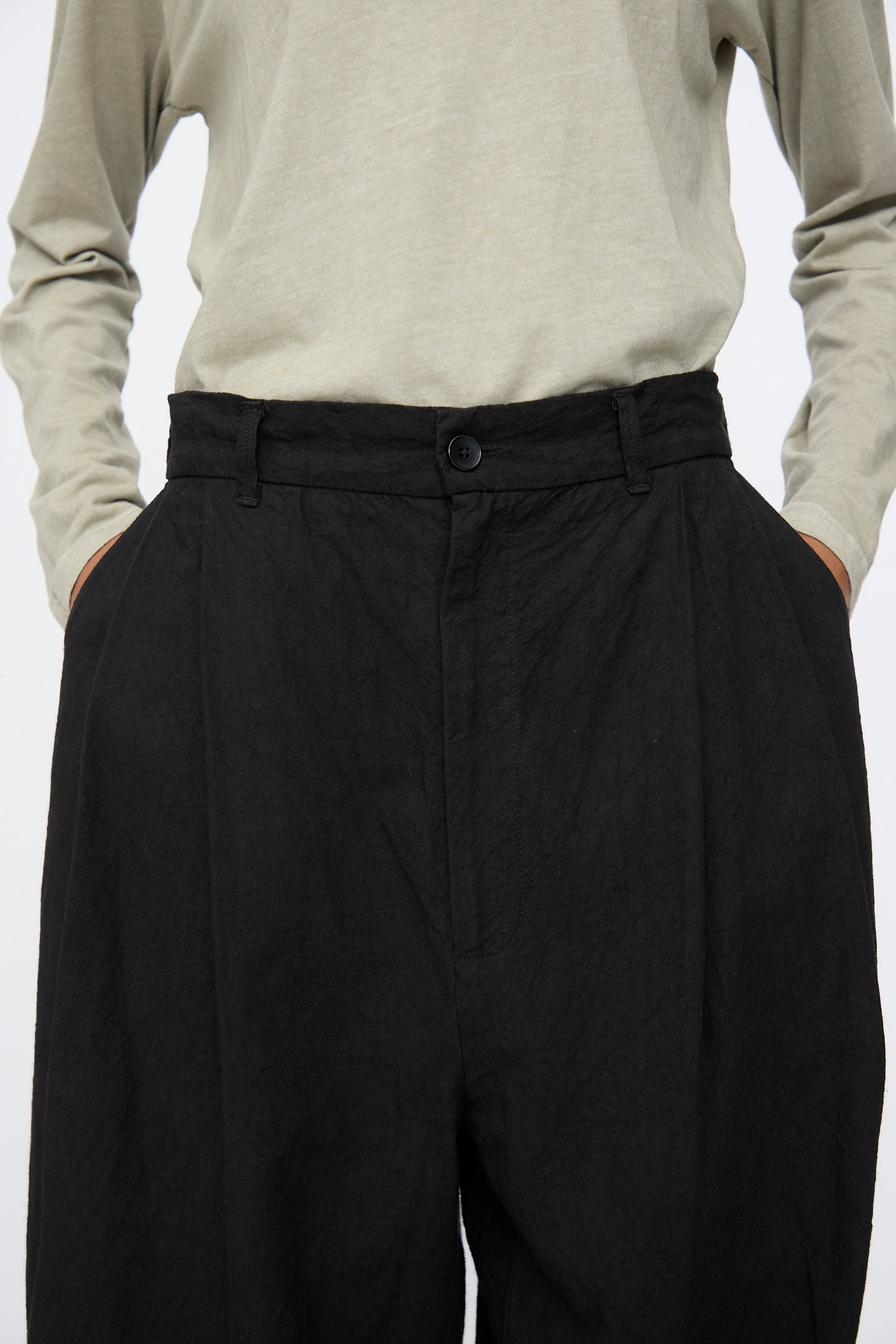 Model wearing Black Crane Crinkled Trousers in Black, paired with a light beige long-sleeve shirt, hands in pockets; shown from shoulders to thighs.