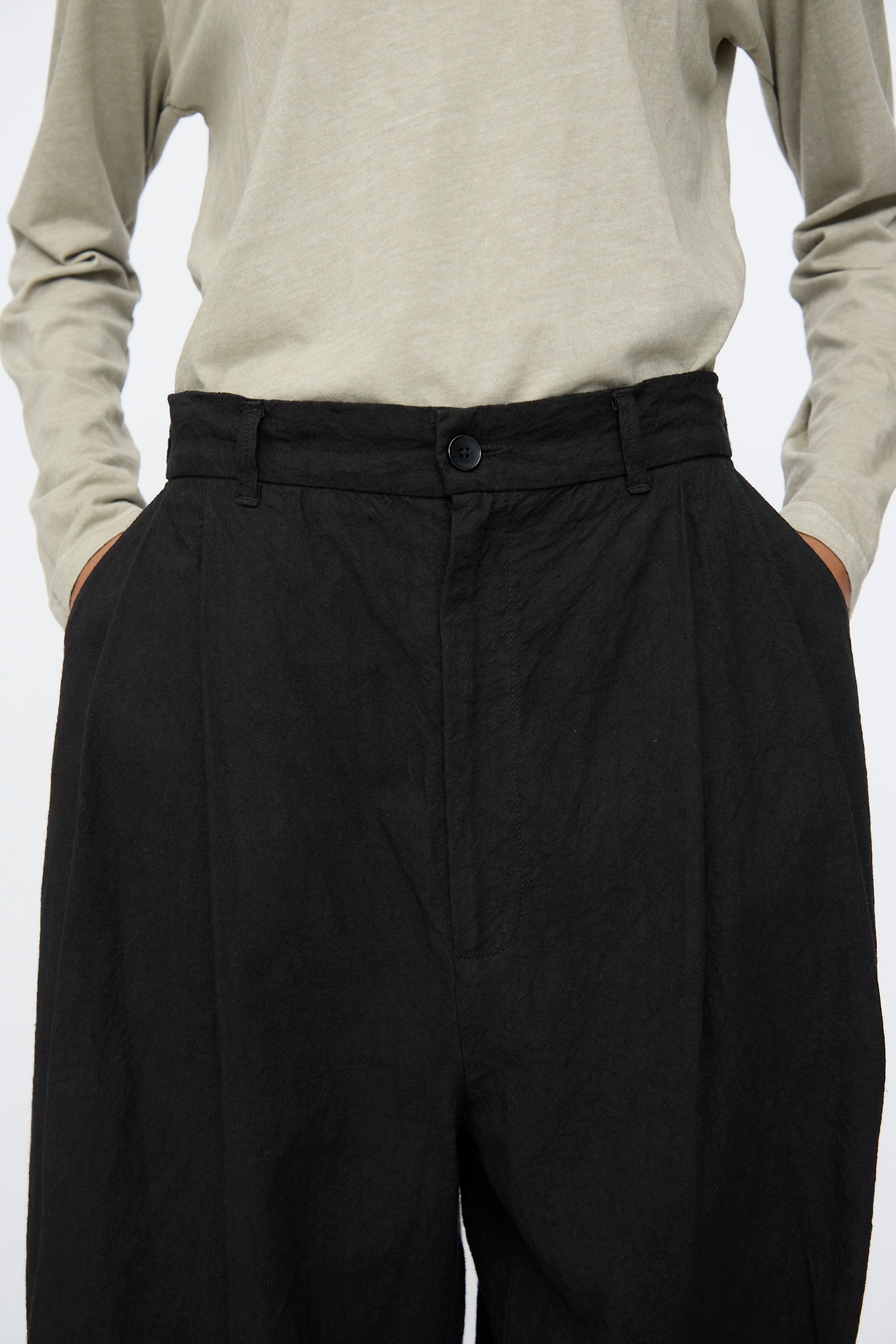 Model wearing Black Crane Crinkled Trousers in Black, paired with a light beige long-sleeve shirt, hands in pockets; shown from shoulders to thighs.