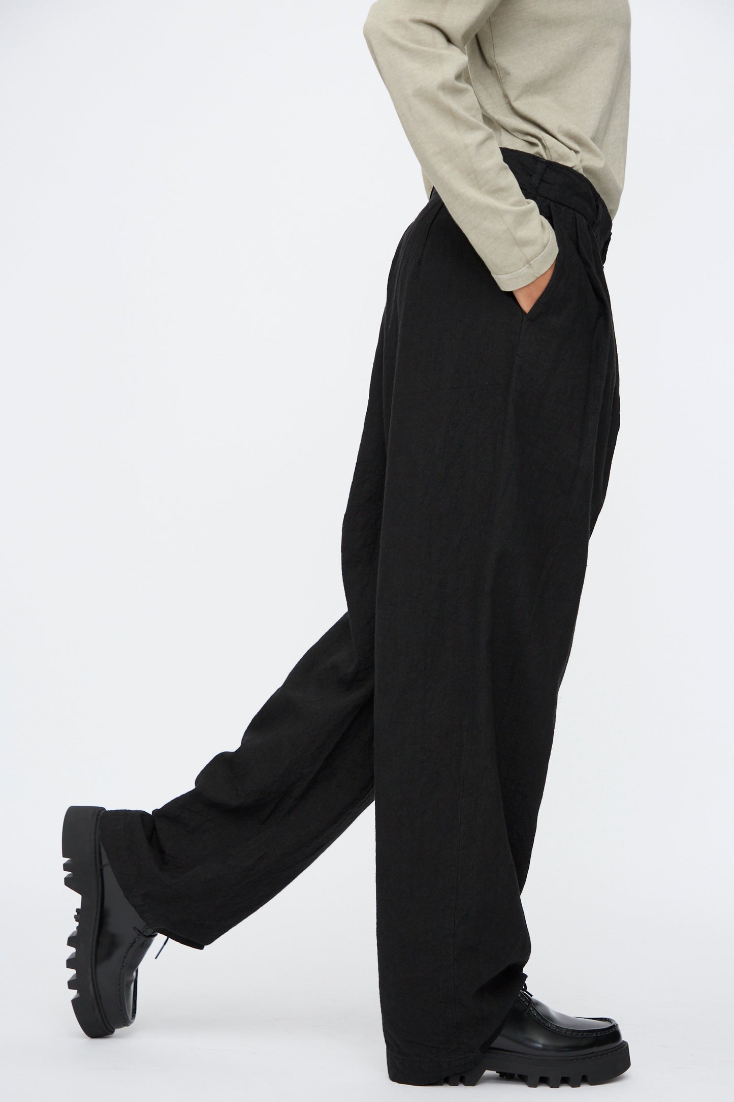 A person wearing Black Crane's Crinkled Trousers in Black, a beige long-sleeve top, and black shoes stands with one leg slightly forward against a plain white background.