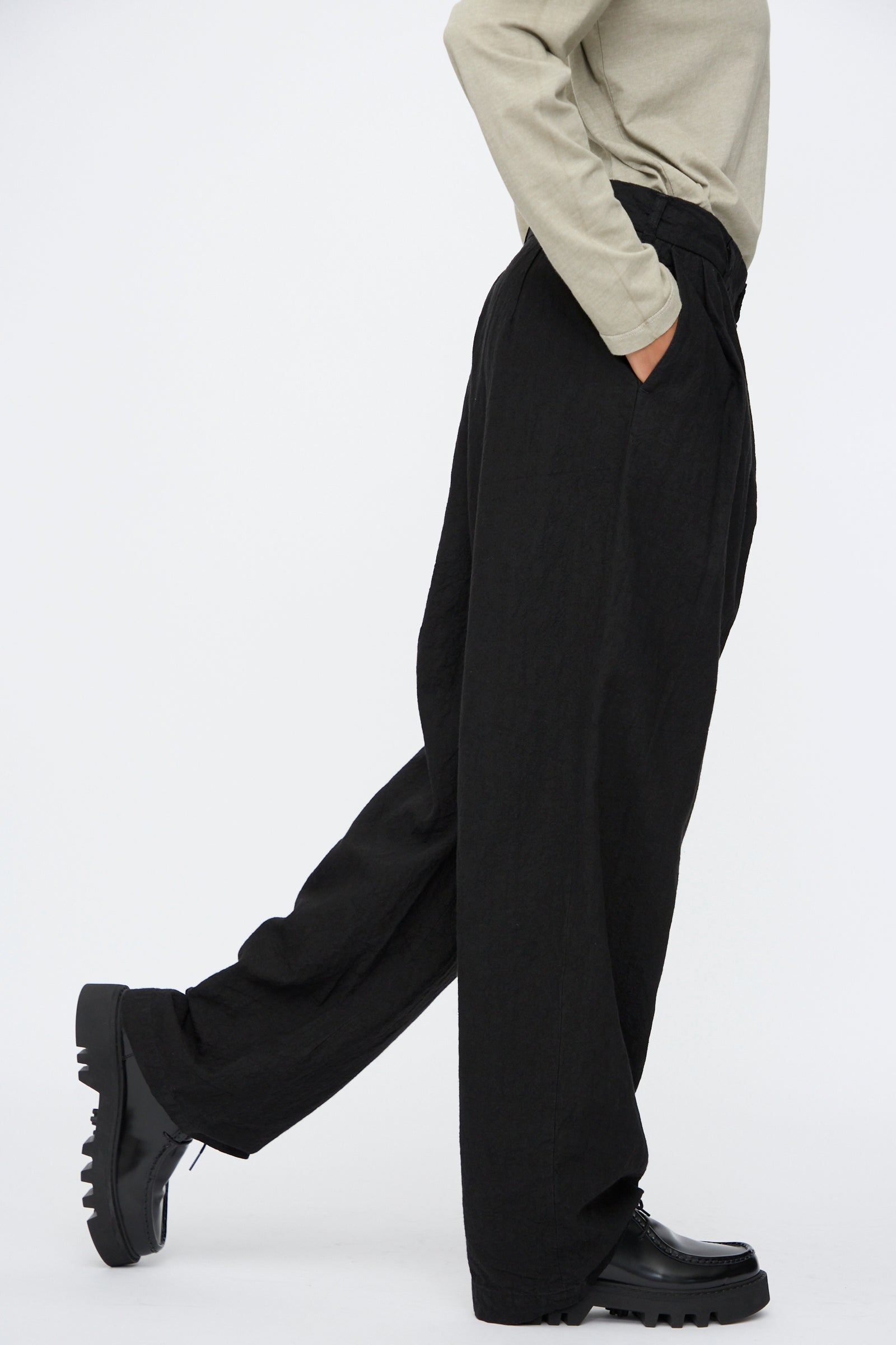 A person wearing Black Crane's Crinkled Trousers in Black, a beige long-sleeve top, and black shoes stands with one leg slightly forward against a plain white background.