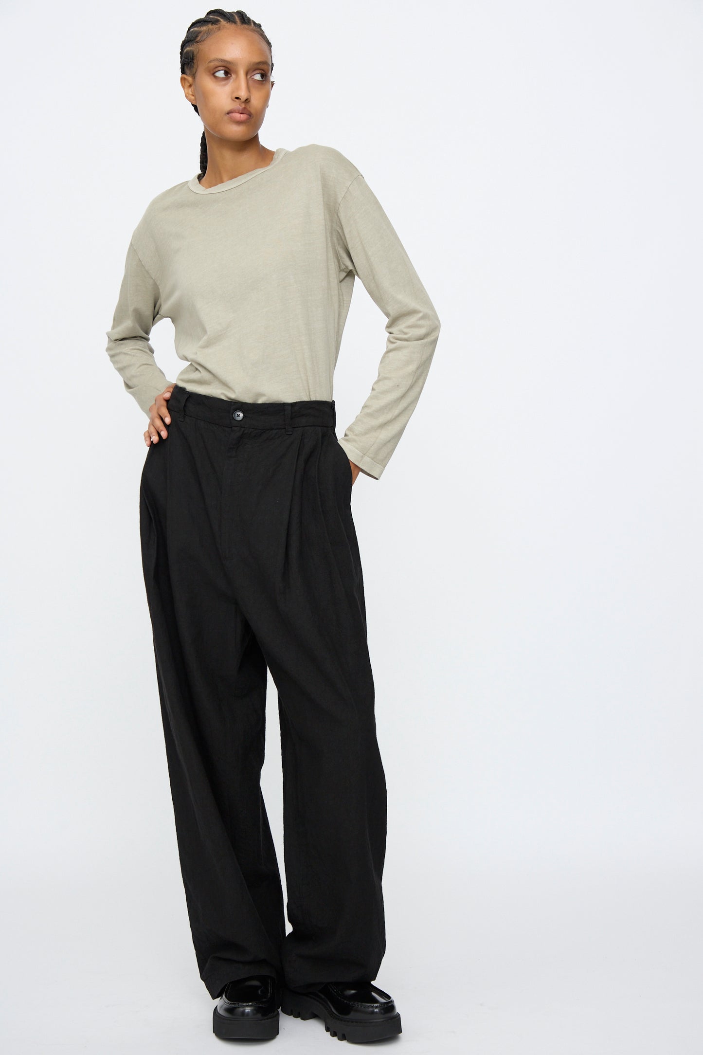 A person stands against a plain background wearing a light beige long-sleeve shirt, Black Crane Crinkled Trousers in Black, and black shoes, with hands in pockets and looking to the side.