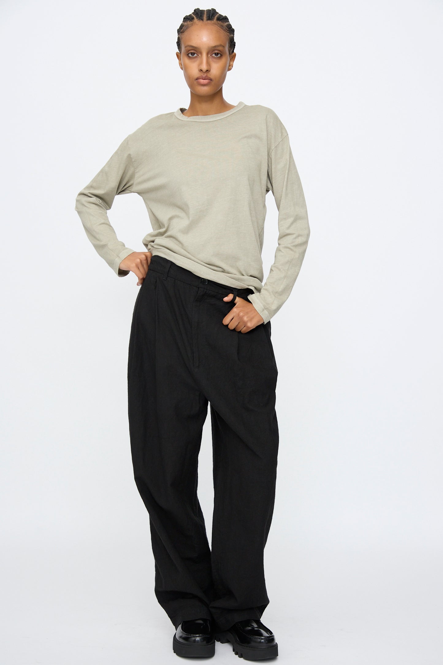 Person stands against a plain background wearing a light beige long-sleeve shirt, Black Crane's Crinkled Trousers in Black, and black shoes, with one hand in their pocket.