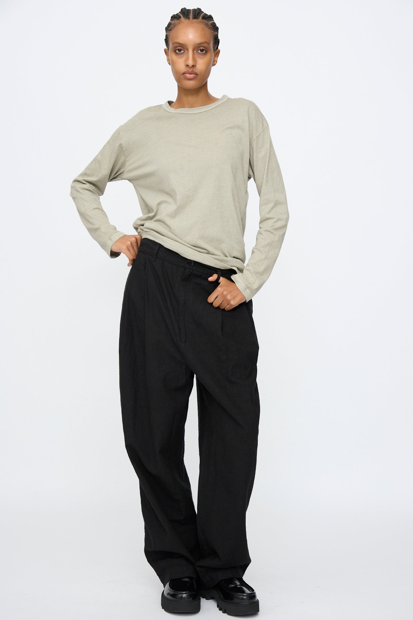Person stands against a plain background wearing a light beige long-sleeve shirt, Black Crane's Crinkled Trousers in Black, and black shoes, with one hand in their pocket.