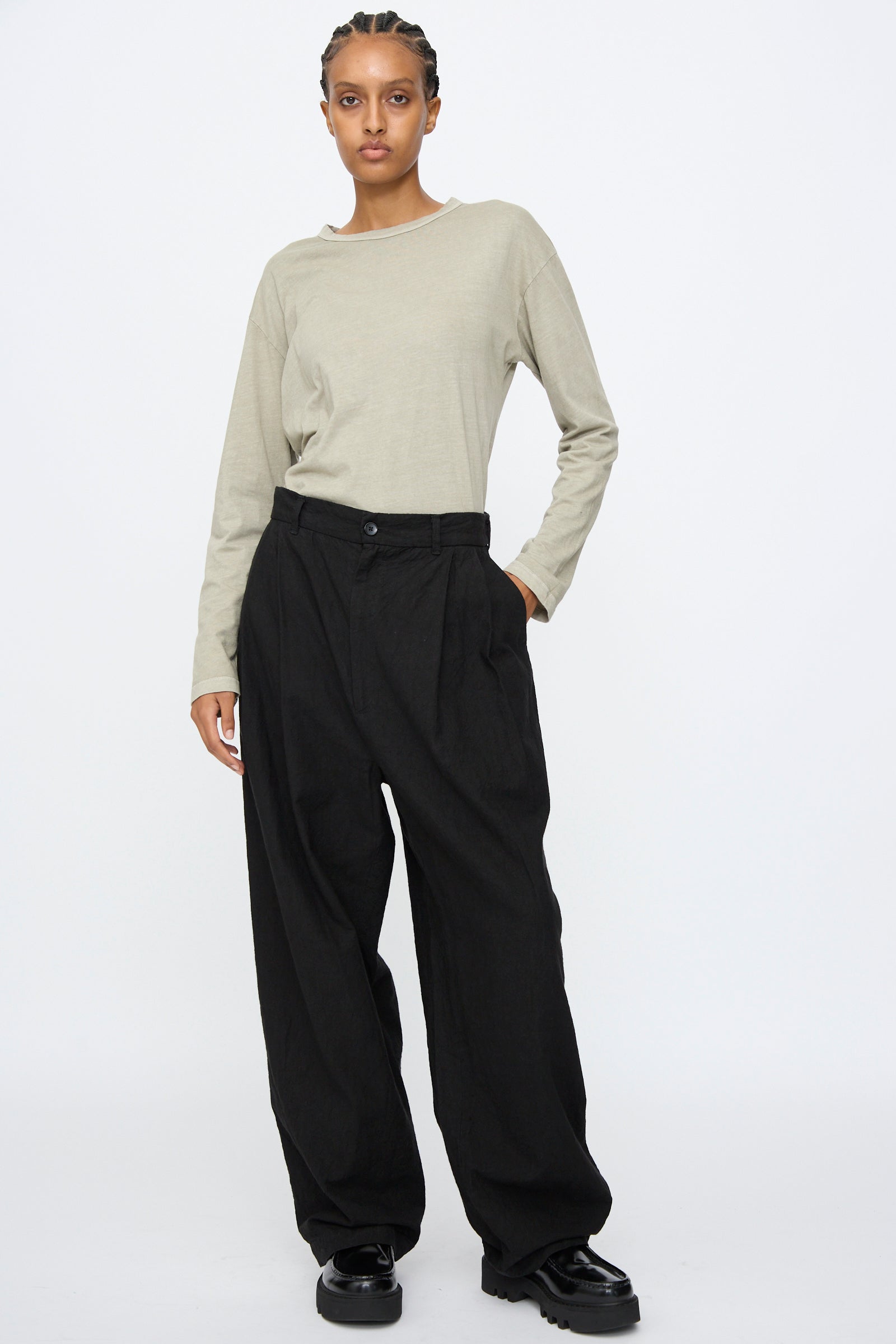 Person standing against a plain background wearing a light beige long-sleeve shirt, Black Crane Crinkled Trousers in Black, and black shoes.