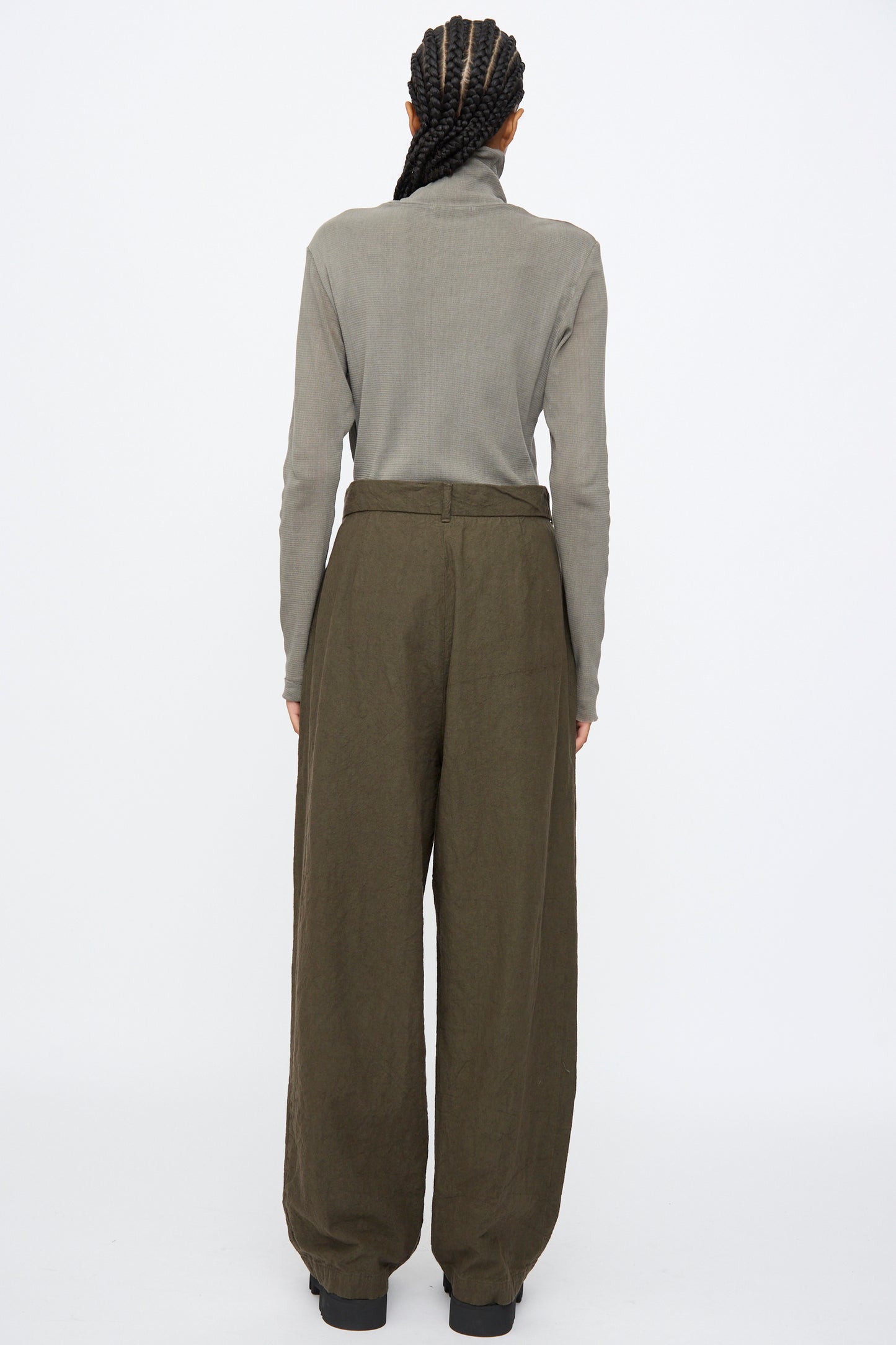 A person with braided hair stands facing away against a white background, wearing a gray turtleneck and Black Crane Crinkled Trousers in Charcoal paired with black shoes.