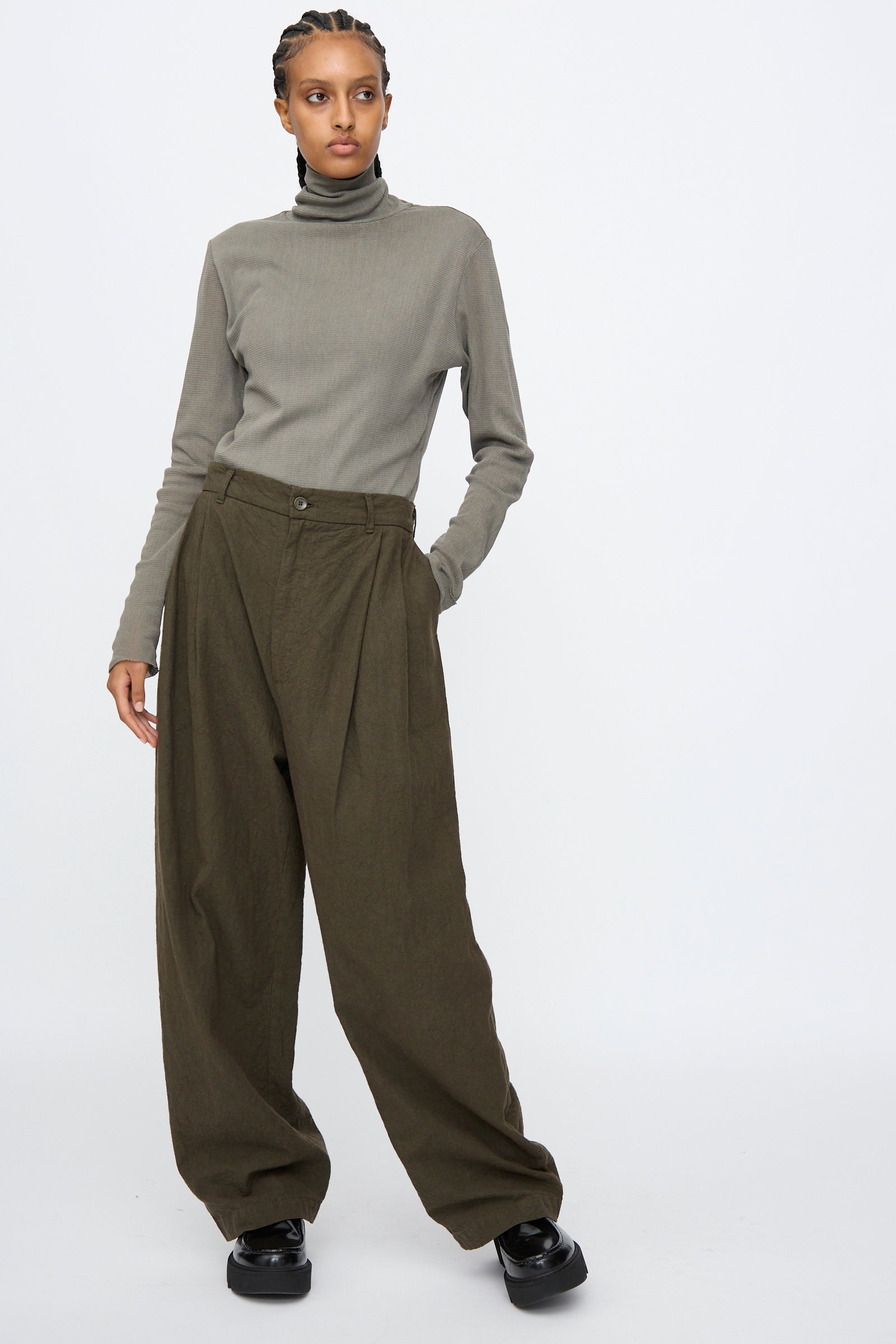 Person wearing a grey turtleneck sweater, Black Crane Crinkled Trousers in Charcoal, and black platform shoes, standing against a plain white background.