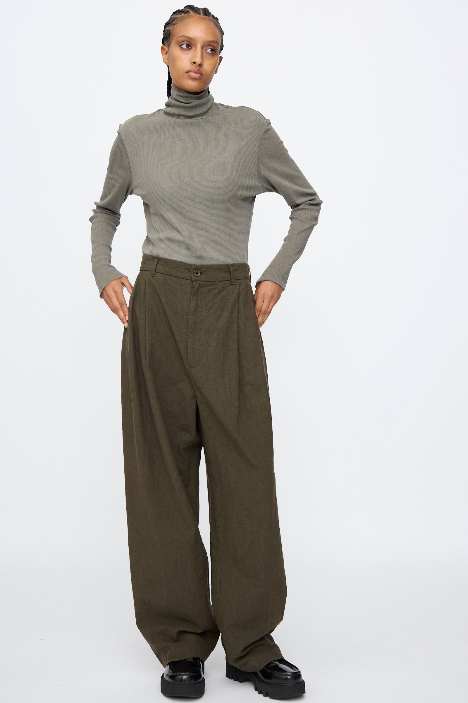 A person stands against a plain background wearing a gray turtleneck sweater, Black Crane Crinkled Trousers in Charcoal, and black shoes.