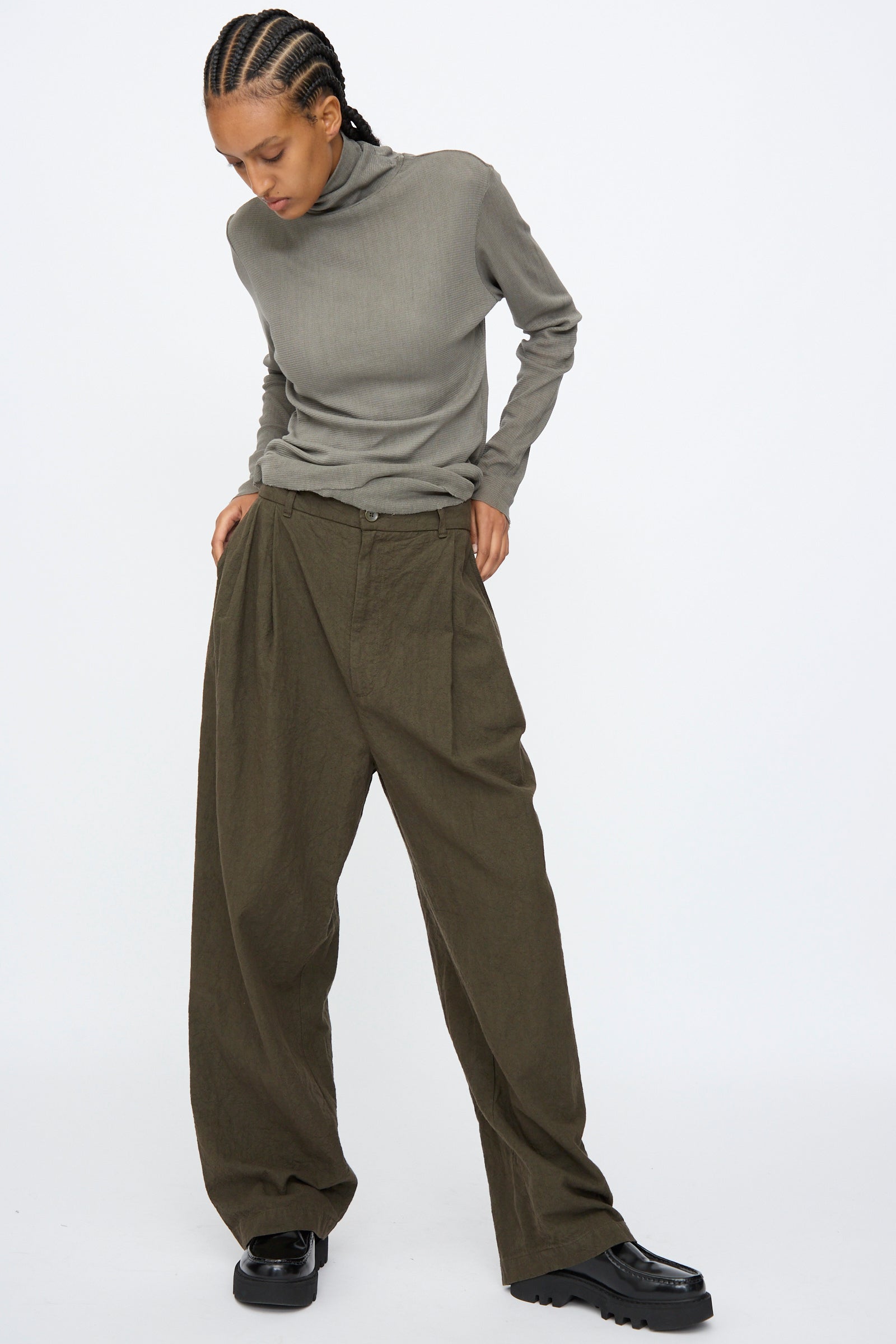 A person stands against a plain light background, looking down with hands in pockets, wearing a gray turtleneck and Black Crane Crinkled Trousers in Charcoal.