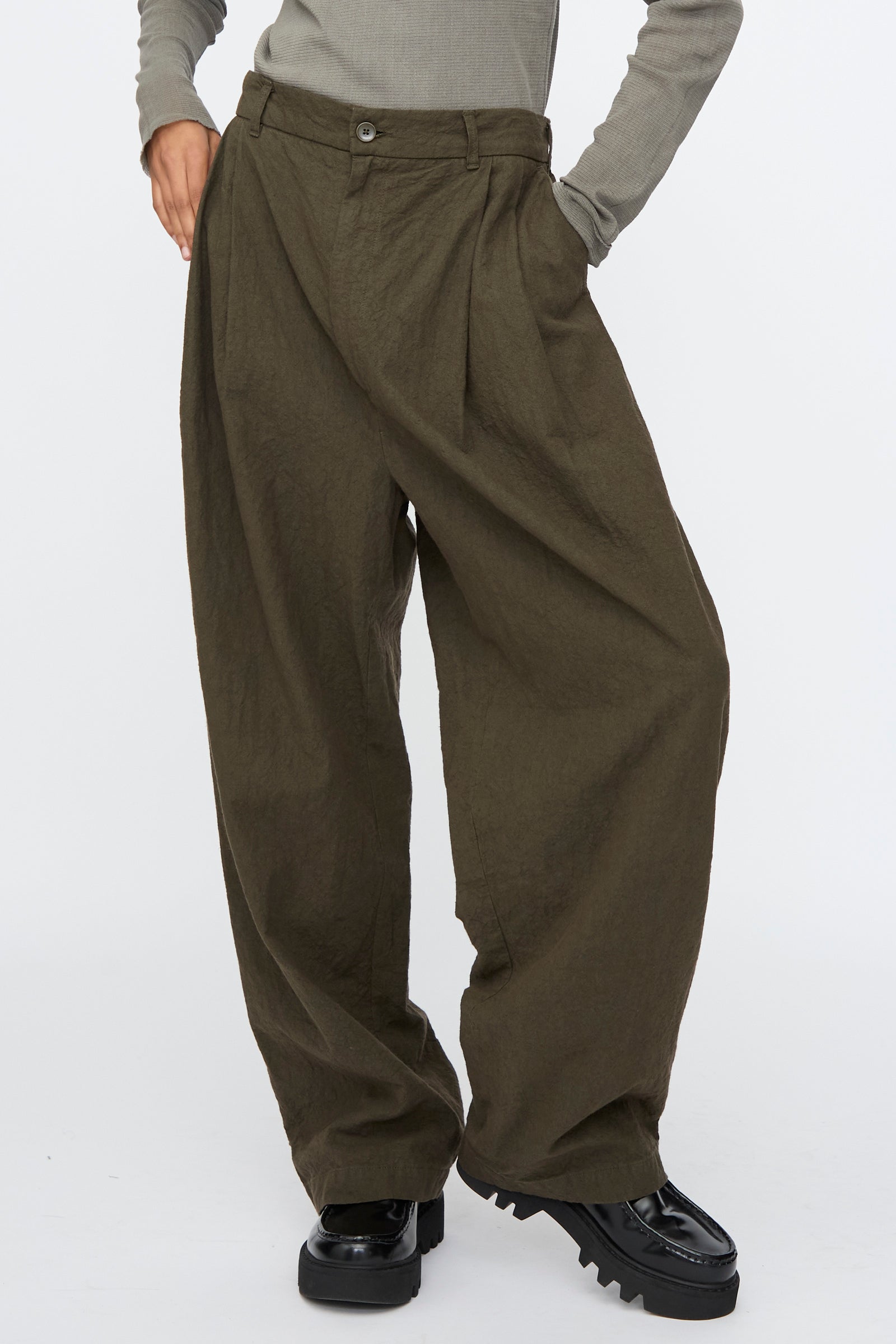 A person wearing Black Crane Crinkled Trousers in Charcoal, a gray long-sleeve top, and black chunky shoes stands against a plain background.