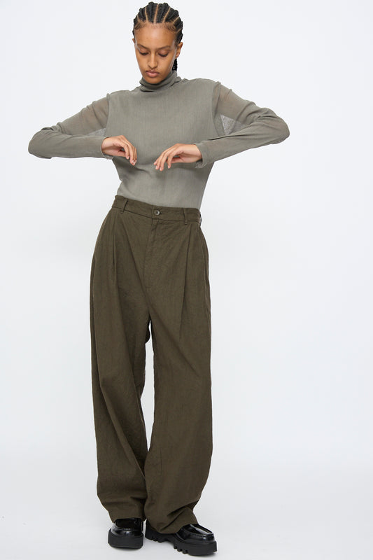 A person wears the Black Crane Crinkled Trousers in Charcoal and a sheer-sleeved gray top, standing against a white background with arms bent and looking down.