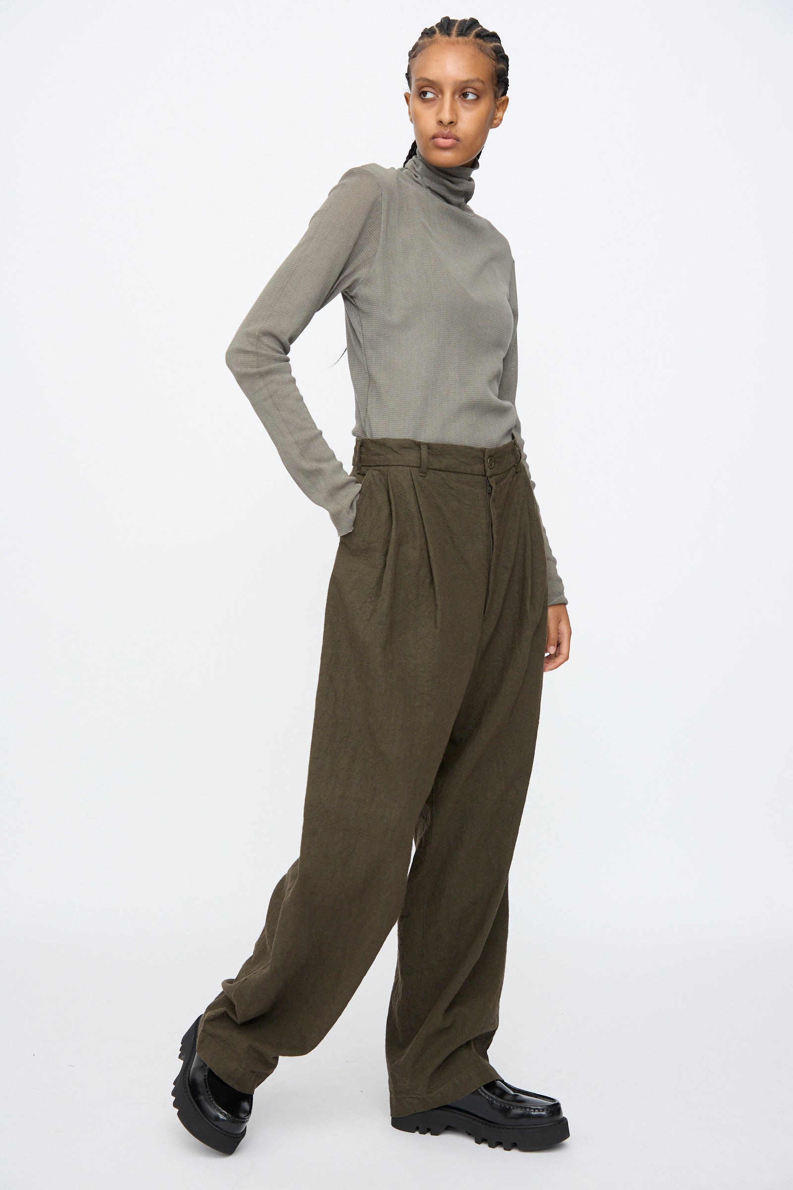 Person stands with one hand in their pocket, wearing a grey long-sleeve turtleneck, Black Crane Crinkled Trousers in Charcoal, and black platform shoes against a plain white background.
