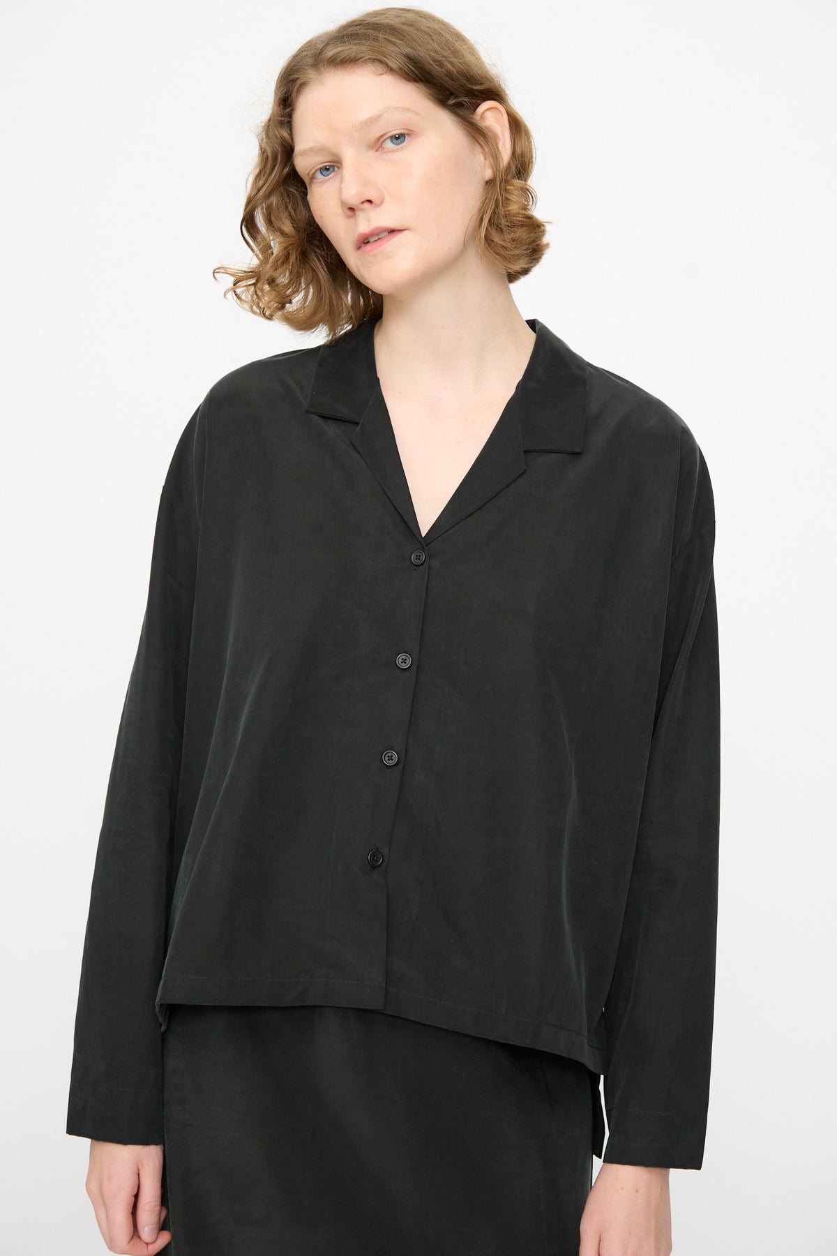 A person with short, wavy hair wears the Black Crane Cropped Shirt in Black—a loose-fitting, boxy long sleeve top made from a soft cotton cupro blend—standing against a plain light background.