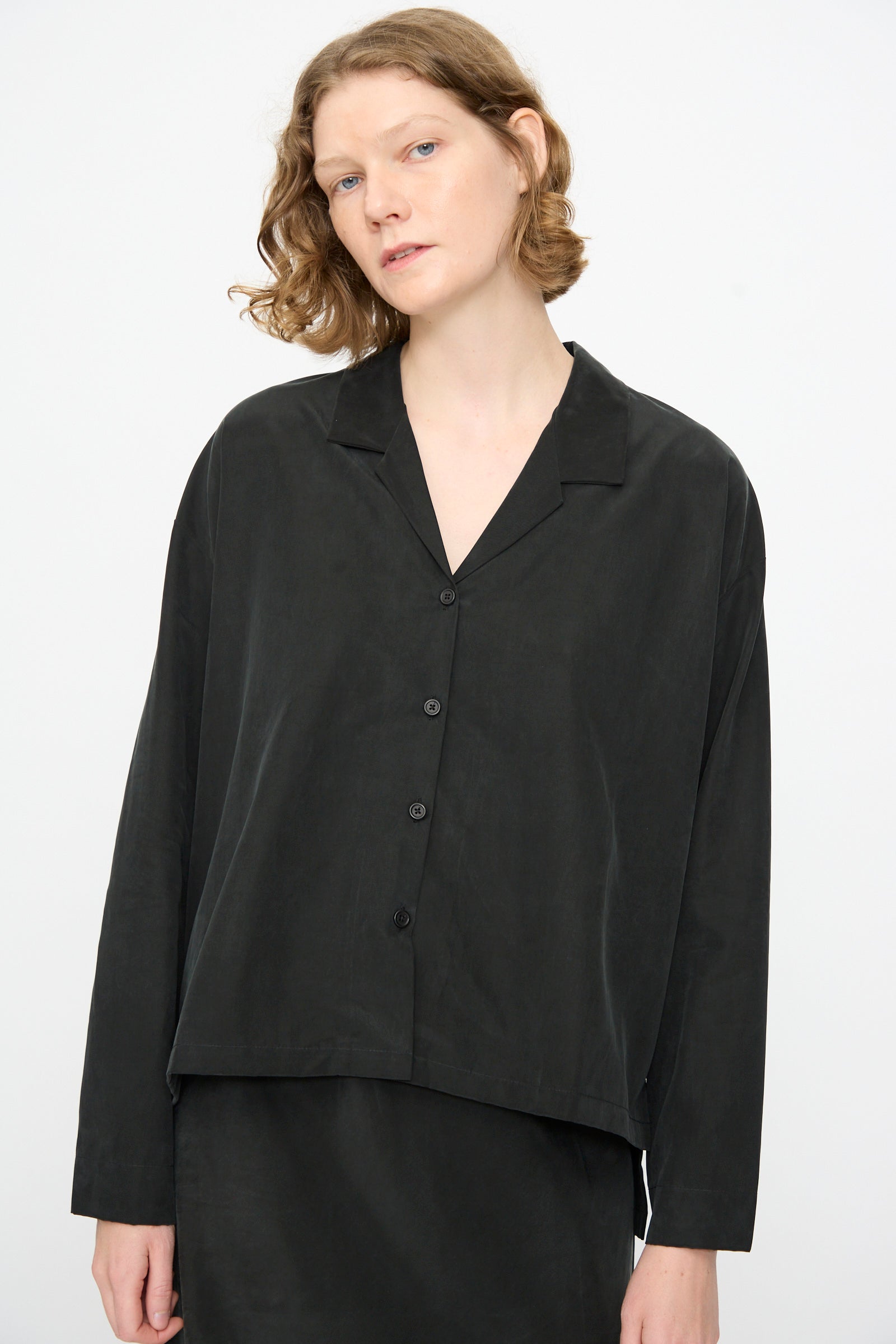 A person with short, wavy hair wears the Black Crane Cropped Shirt in Black—a loose-fitting, boxy long sleeve top made from a soft cotton cupro blend—standing against a plain light background.