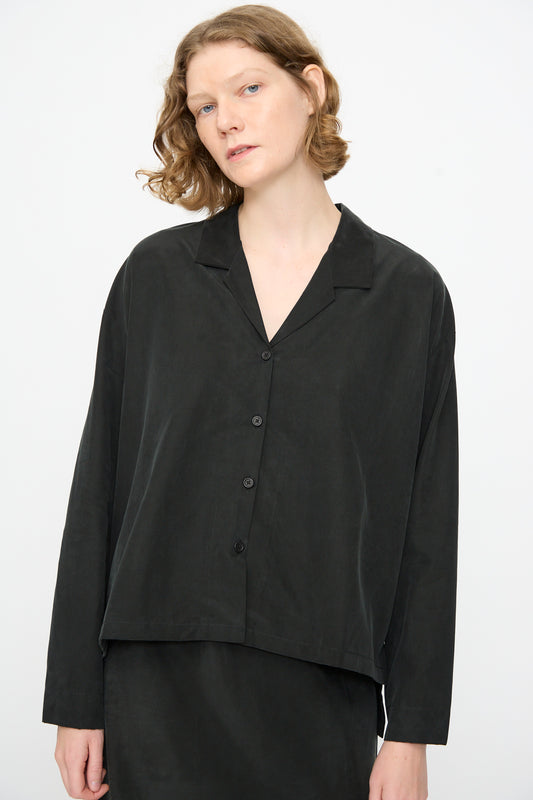 A person with short, wavy hair wears the Black Crane Cropped Shirt in Black—a loose-fitting, boxy long sleeve top made from a soft cotton cupro blend—standing against a plain light background.