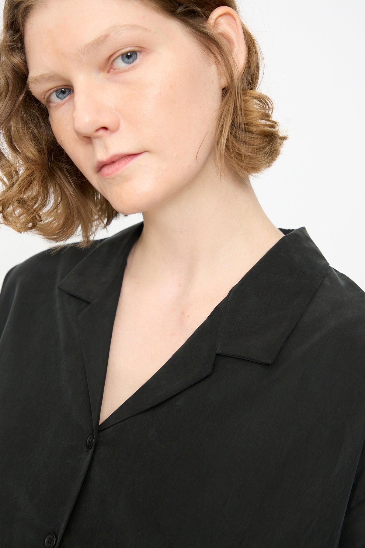 A person with short wavy light brown hair wears the Black Crane Cropped Shirt in Black, a long sleeve collared top crafted from a cotton cupro blend, and looks into the camera against a plain white background.
