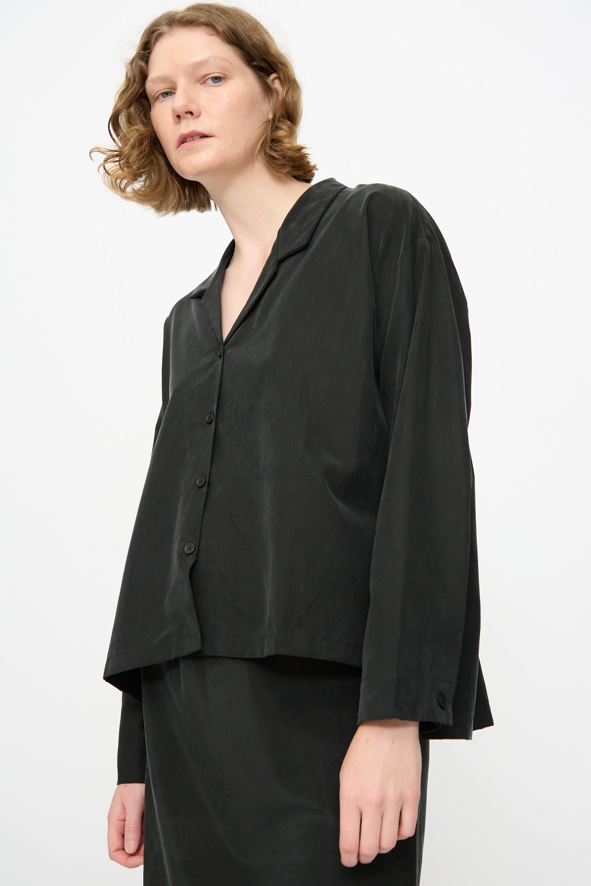 A person wearing the Black Crane Cropped Shirt in Black—a loose-fitting, boxy, long-sleeve button-up crafted from a soft cotton cupro blend—paired with a matching black skirt, standing against a plain white background.