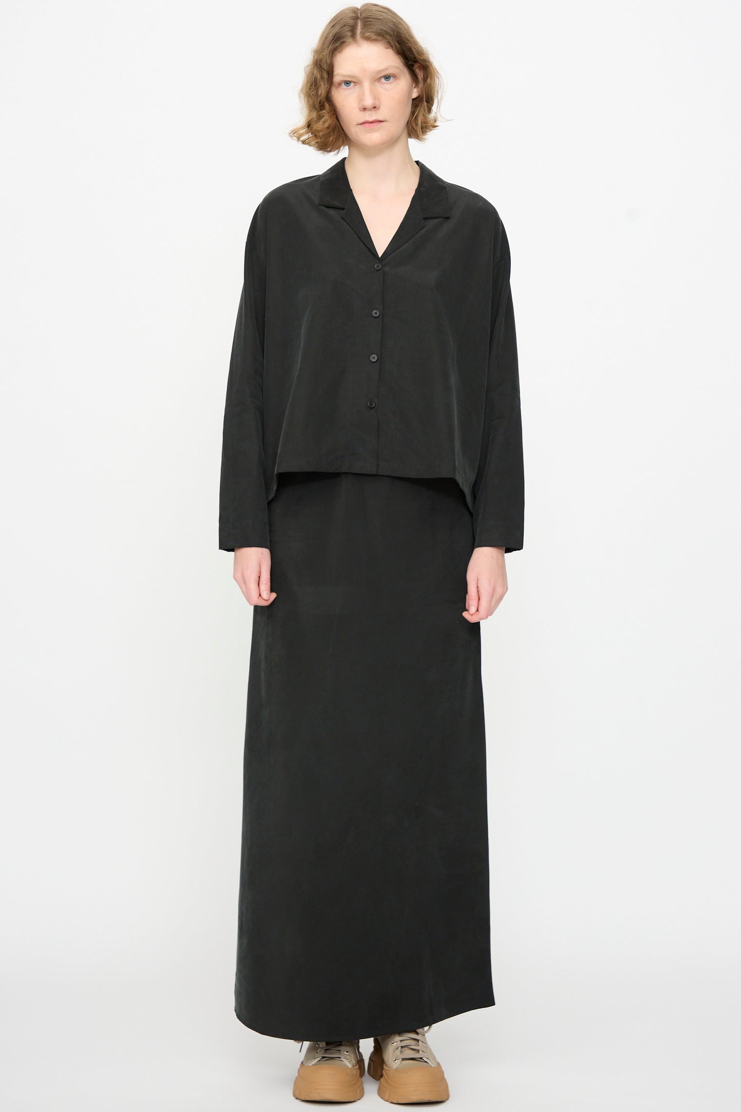 A person stands against a plain white background, facing forward with a neutral expression, wearing the Black Crane Cropped Shirt in Black—a loose, boxy-fit cotton cupro blend button-up top—paired with a long black skirt and beige shoes.