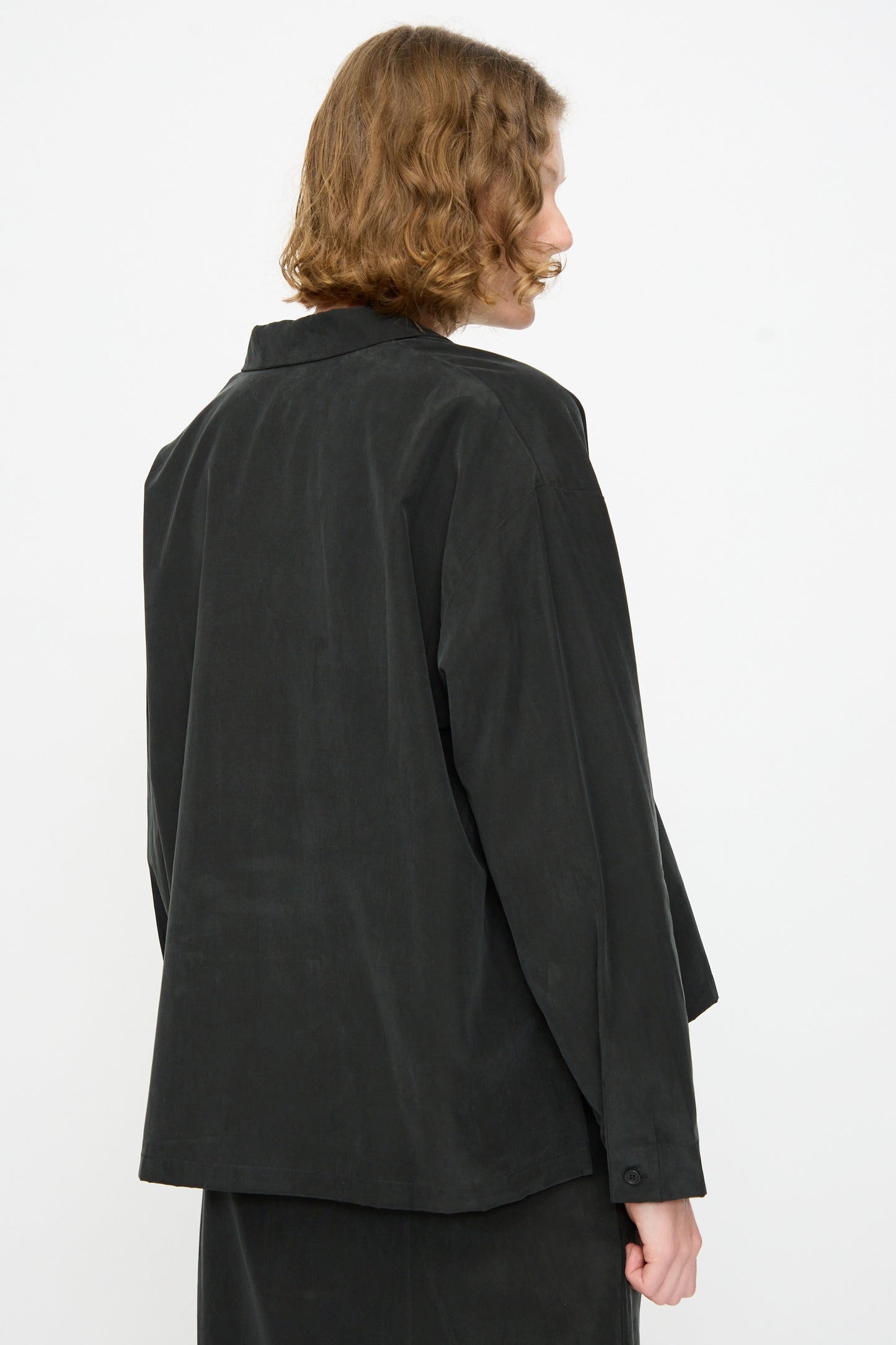 A person with short, wavy brown hair is shown from the back against a plain white background, wearing the Black Crane Cropped Shirt in Black—a loose-fitting, boxy jacket-style top.