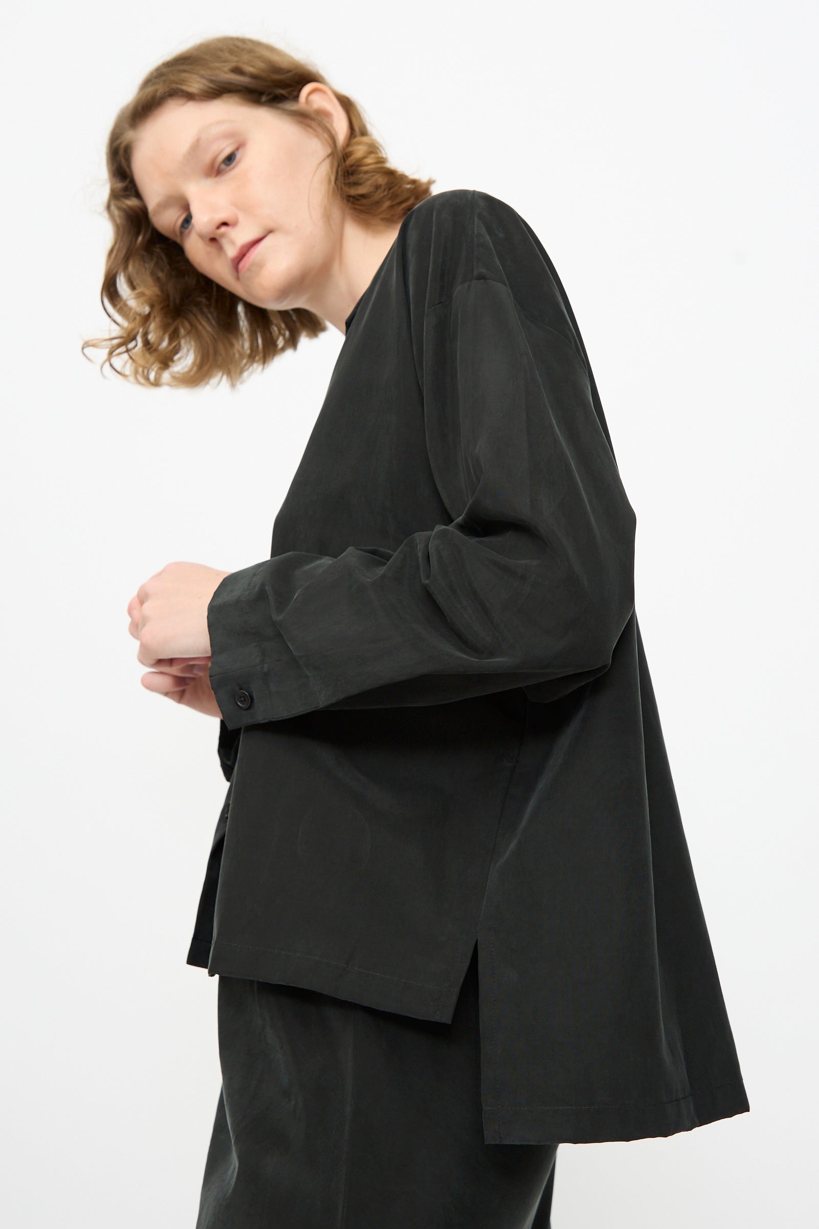 A person with shoulder-length light brown hair is wearing the Black Crane Cropped Shirt in Black—a loose, boxy-fit, cropped long sleeve shirt—paired with matching pants crafted from a soft cotton cupro blend, standing against a plain white background.