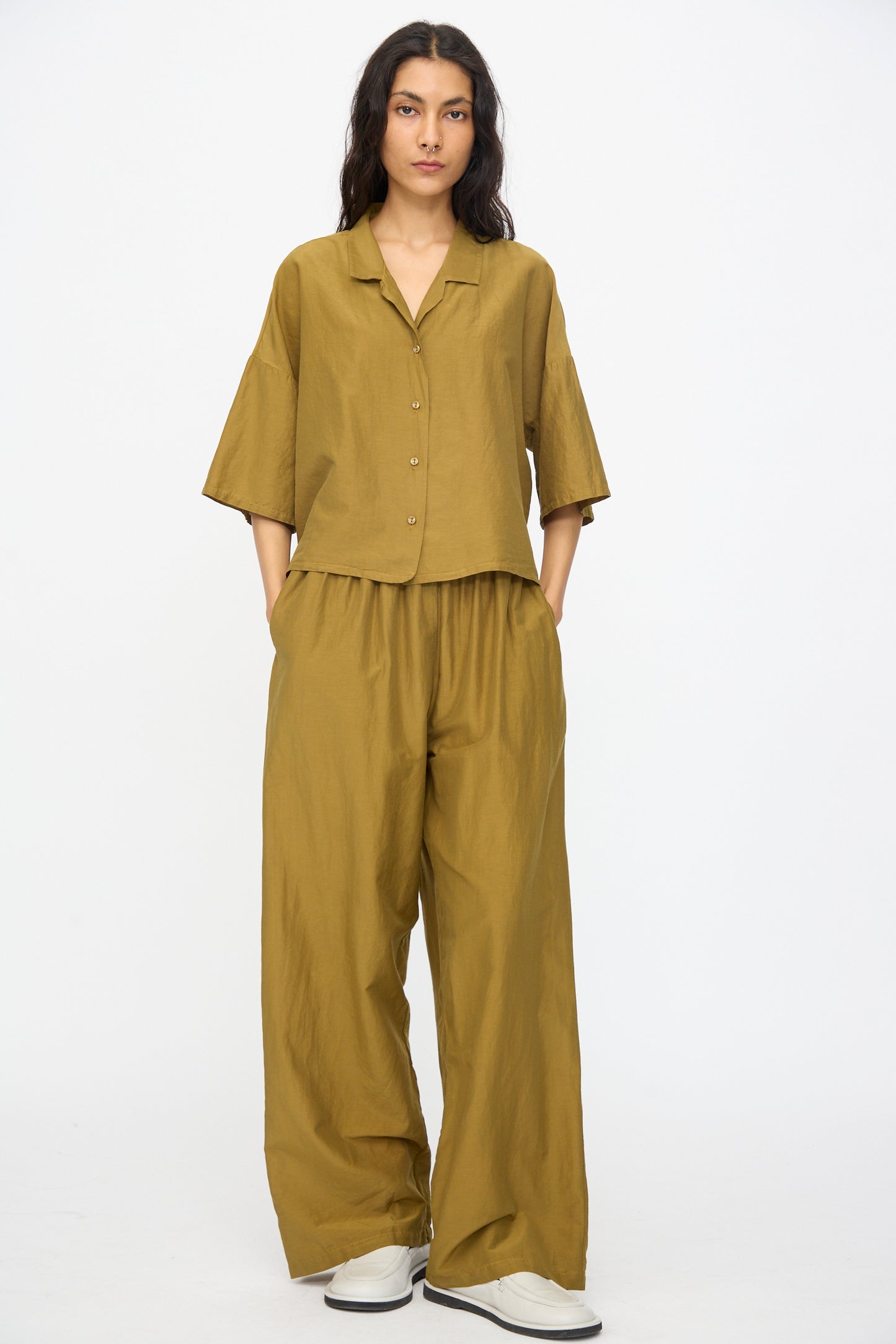 Person standing against a white background, wearing the Black Crane Cropped Shirt in Dark Olive. The shirt is short sleeve with a boxy fit and is paired with wide-leg pants and white slip-on shoes. Hands are in their pockets.