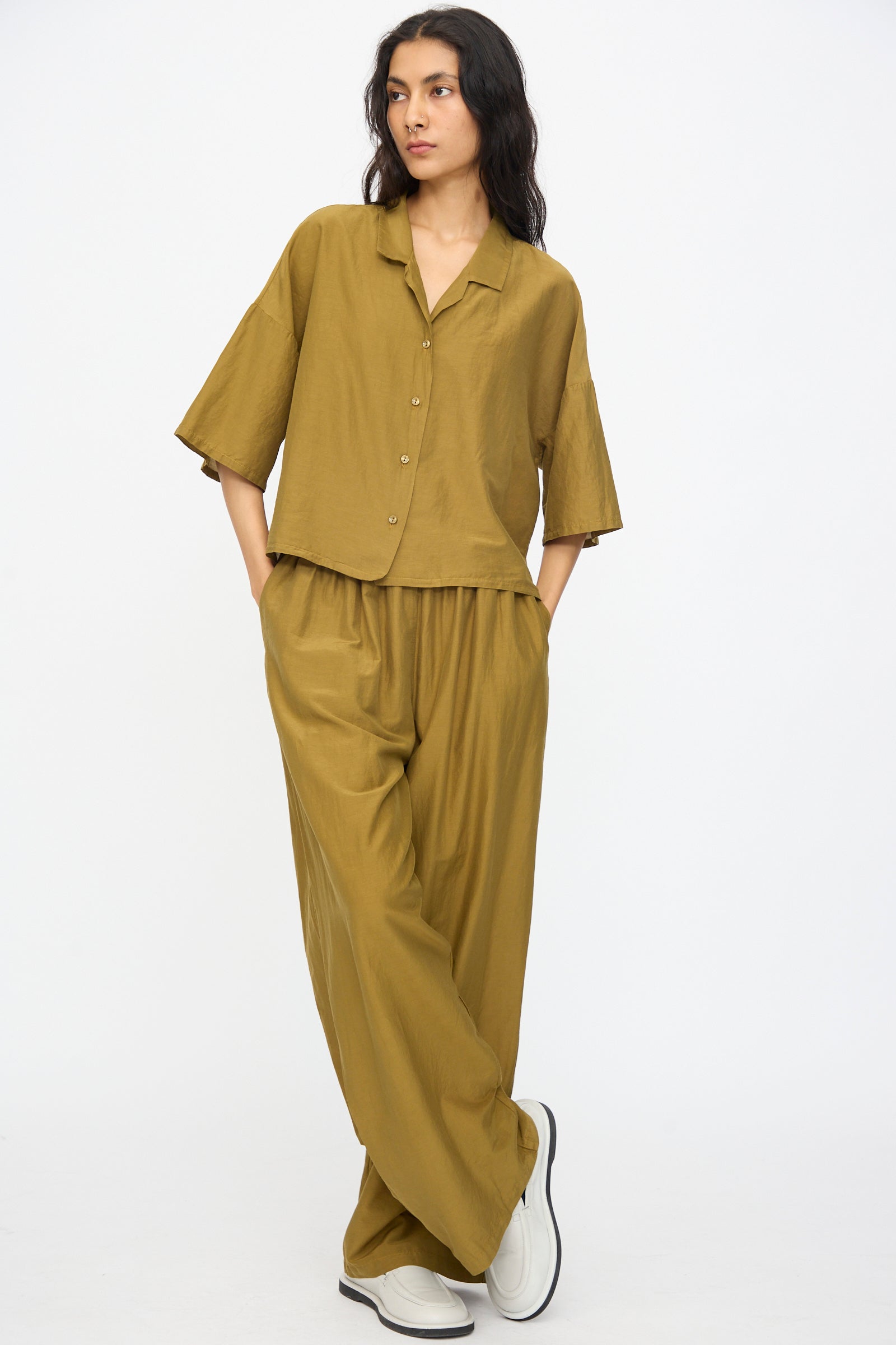 A person wearing the Black Crane Cropped Shirt in Dark Olive with hands in pockets and white slip-on shoes, standing against a plain background.