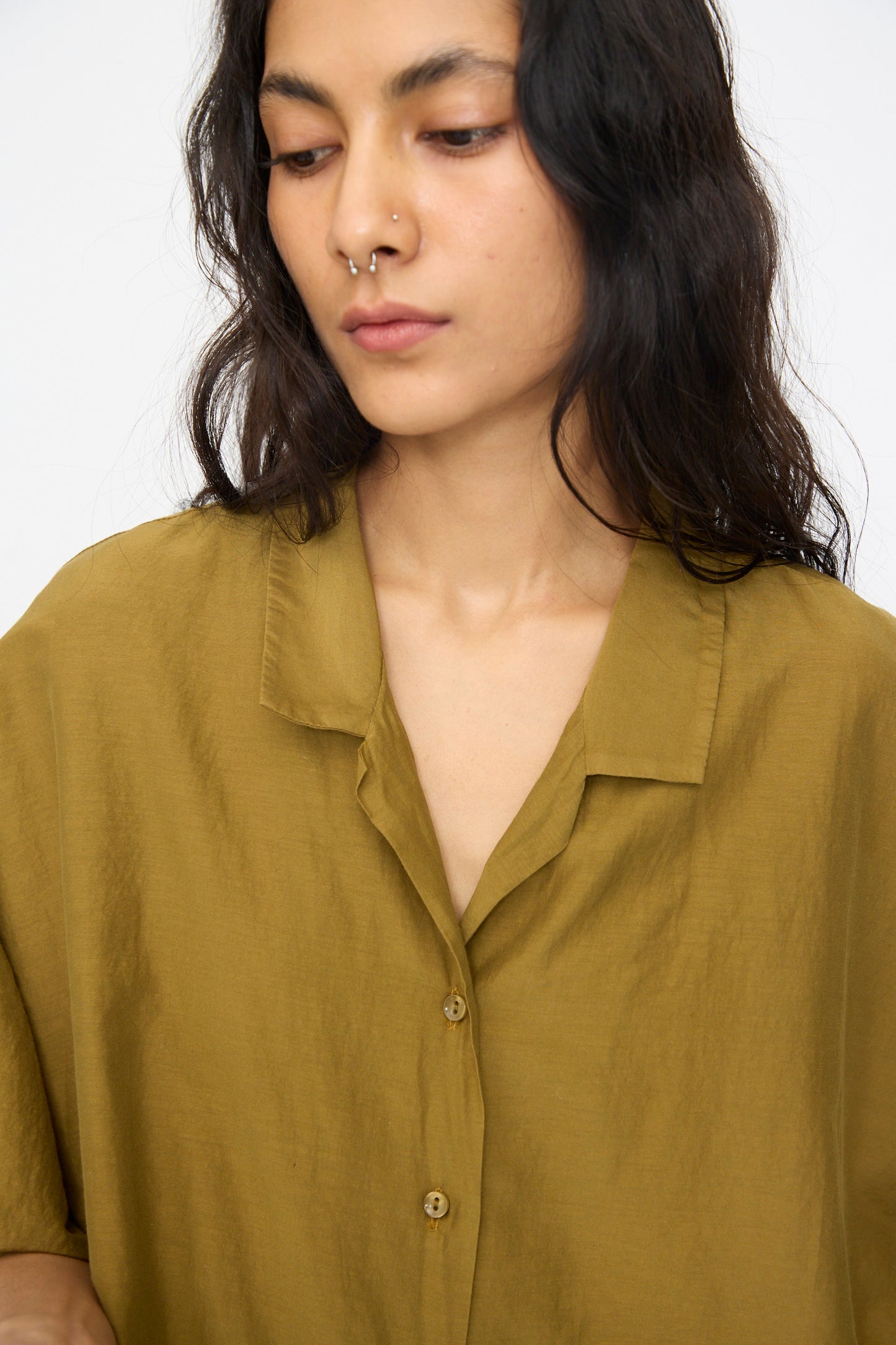 Person with long dark hair and a septum piercing wears the Black Crane Cropped Shirt in Dark Olive, a boxy-fit button-up made from a soft cotton-silk blend, looking down with a neutral expression.