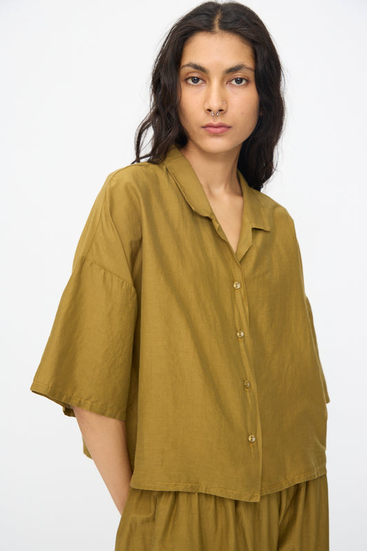 Person with long dark hair wearing the Black Crane Cropped Shirt in Dark Olive, standing against a plain light background.