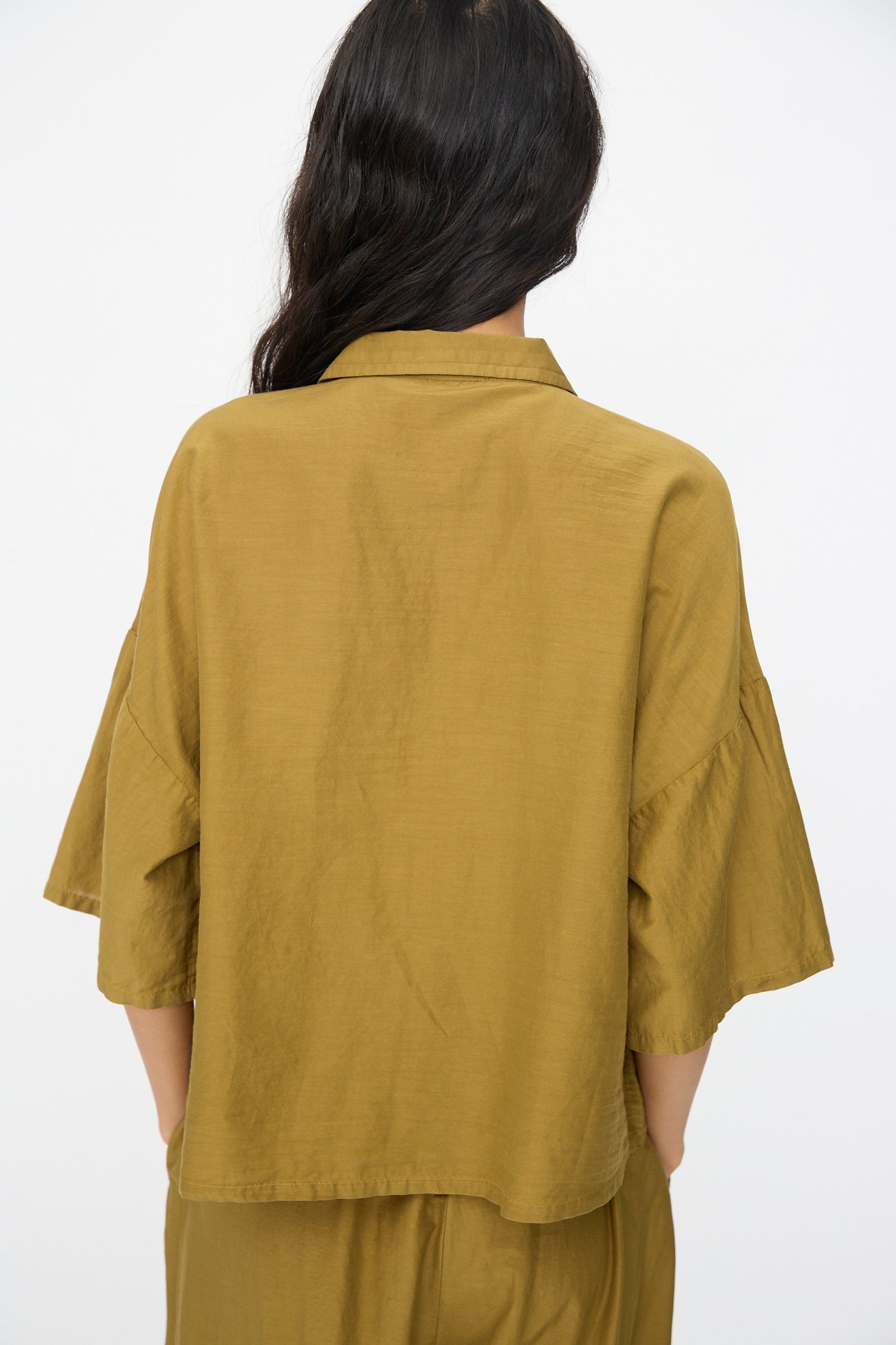 A person with long dark hair is seen from behind wearing the Black Crane Cropped Shirt in Dark Olive, featuring a boxy fit and wide sleeves, paired with matching pants, all on a plain light background.