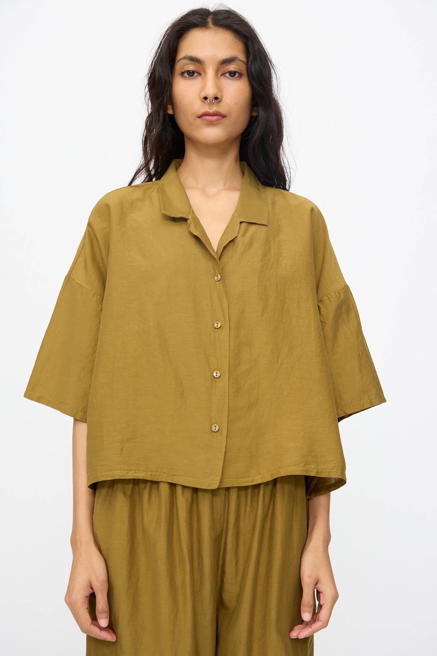 A person wearing Black Crane's Cropped Shirt in Dark Olive, featuring a boxy fit and short sleeves, stands against a plain white background with matching pants in a soft cotton silk blend.