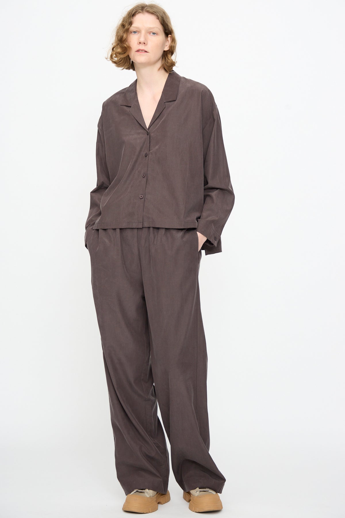 A person stands against a white background, wearing the Black Crane Cropped Shirt in Dark Plum—a loose-fitting, boxy collared button-up crafted from a cotton cupro blend—paired with wide-legged pants and tan shoes.