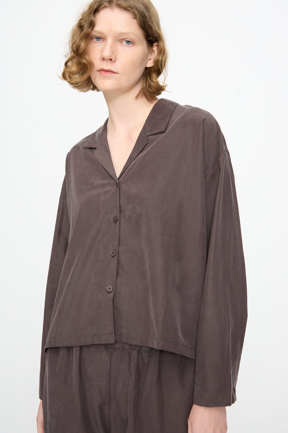 A person wearing the Black Crane Cropped Shirt in Dark Plum, a loose-fitting, boxy long sleeve shirt with a collar crafted from a soft cotton cupro blend and paired with matching pants, stands against a plain white background. The relaxed silhouette of the outfit enhances its modern appeal.