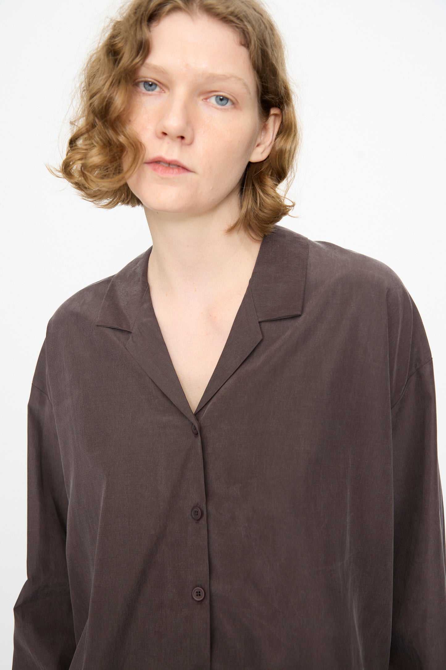 A person with short, wavy, light brown hair is wearing the Black Crane Cropped Shirt in Dark Plum—a boxy-fit, long sleeve button-up crafted from a soft cotton cupro blend—while looking at the camera against a plain white background.