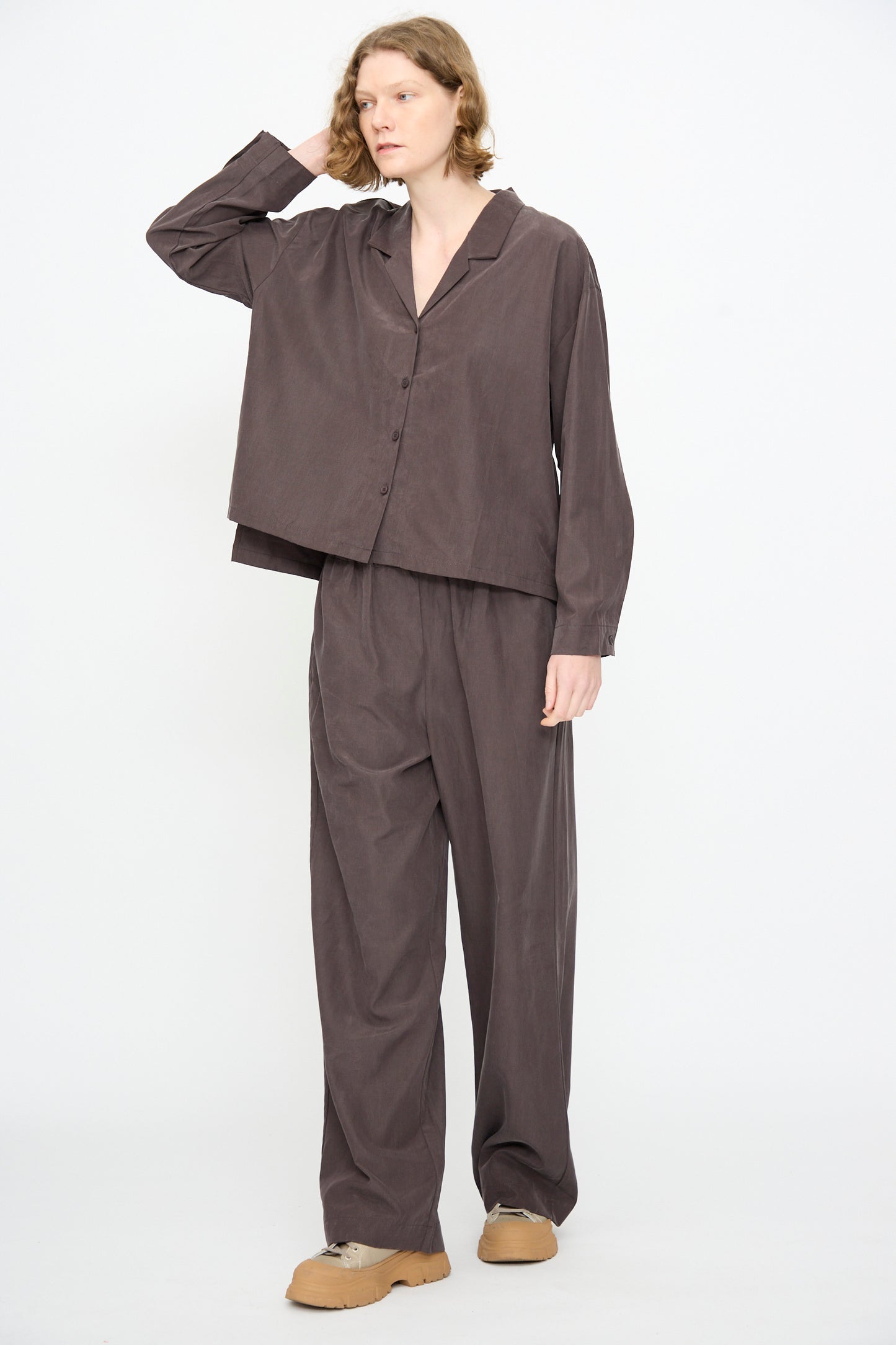 A person stands against a white background, wearing the Black Crane Cropped Shirt in Dark Plum—a loose-fitting, boxy button-up—paired with matching wide-leg pants crafted from a soft cotton cupro blend and tan shoes, with one hand touching their hair.