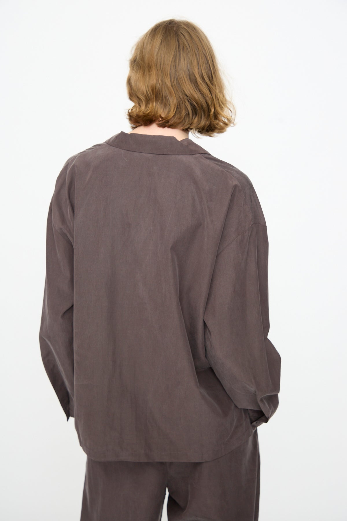A person with shoulder-length light brown hair is seen from the back against a plain white background, wearing Black Crane’s Cropped Shirt in Dark Plum—a loose-fitting, boxy long-sleeved top crafted from a soft cotton cupro blend.